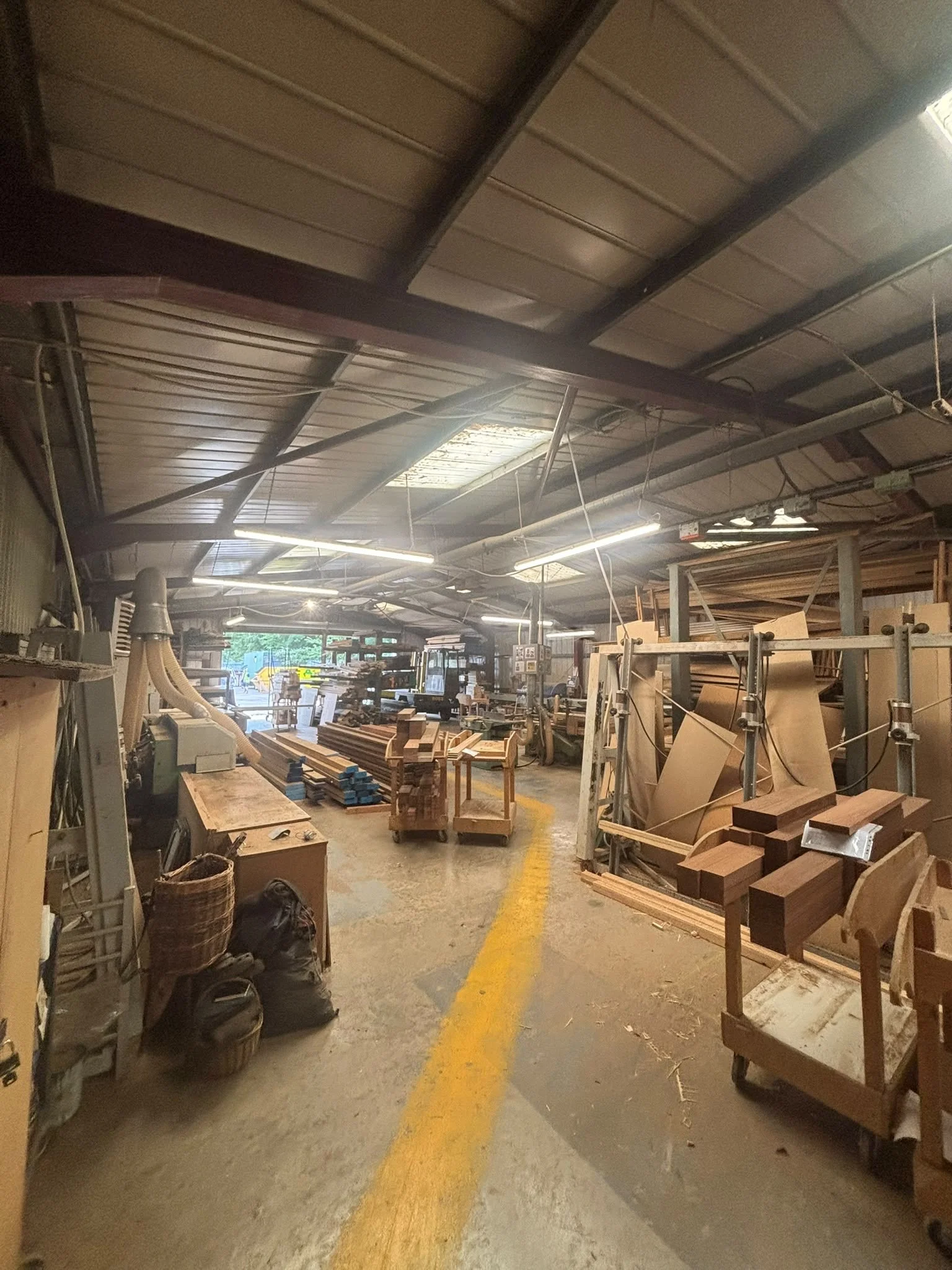 Interior of a woodworking workshop with various woodworking tools, wooden pieces, carts, and a yellow line on the concrete floor.