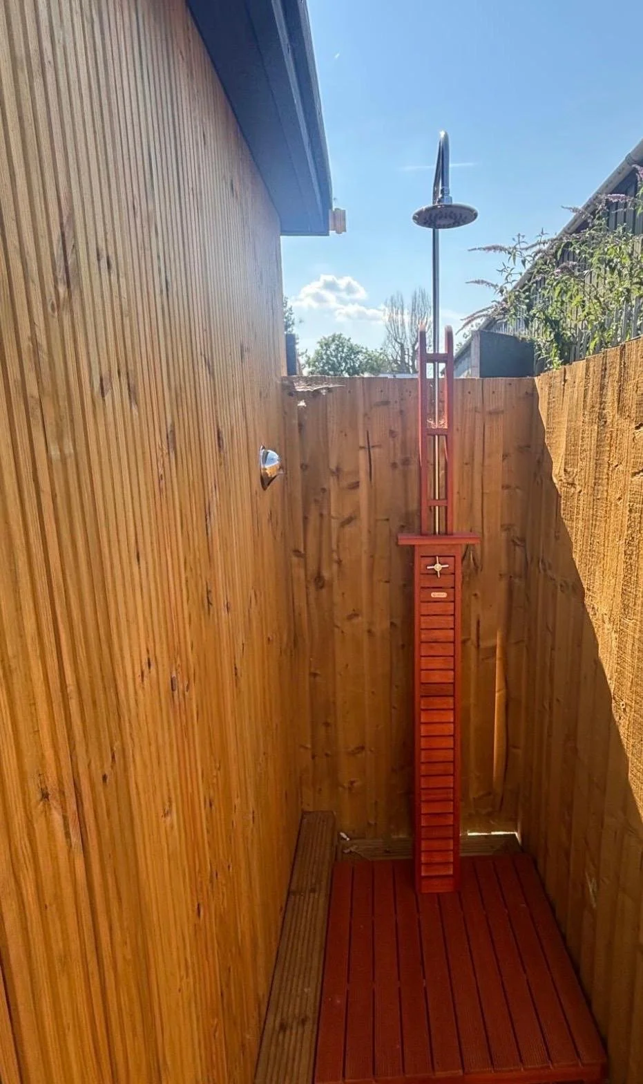 An outdoor shower with a rain head, mounted on a wooden wall, in a small fenced shower area with a wooden floor.