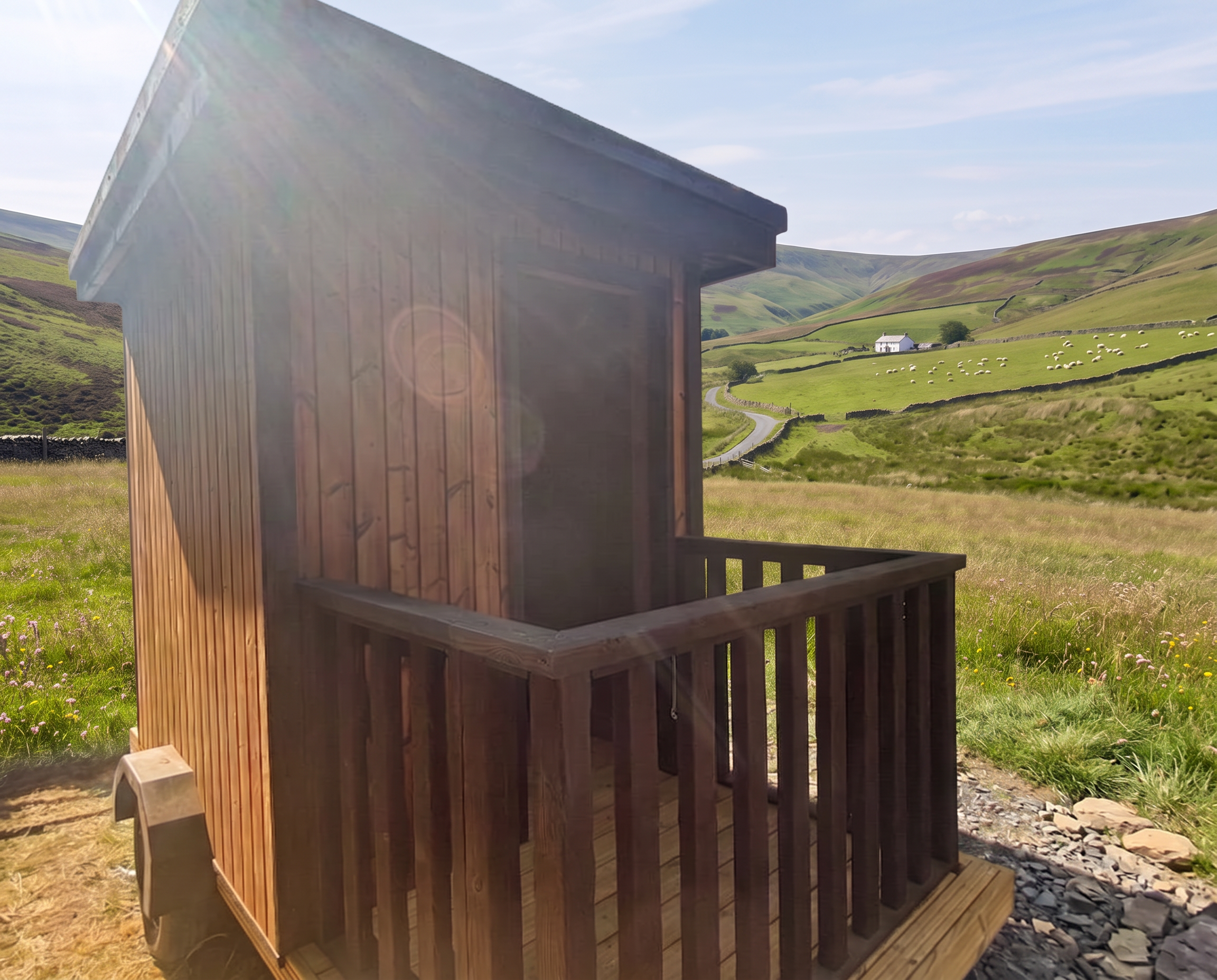 A small wooden cabin with a balcony in a green rural area with hills and a white house in the background under blue sky with some clouds.
