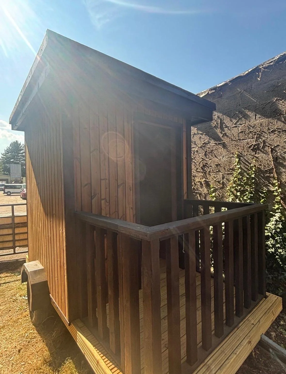 A small wooden structure with a slanted roof and a screened opening, featuring a balcony with wooden railing, situated outdoors in bright sunlight.