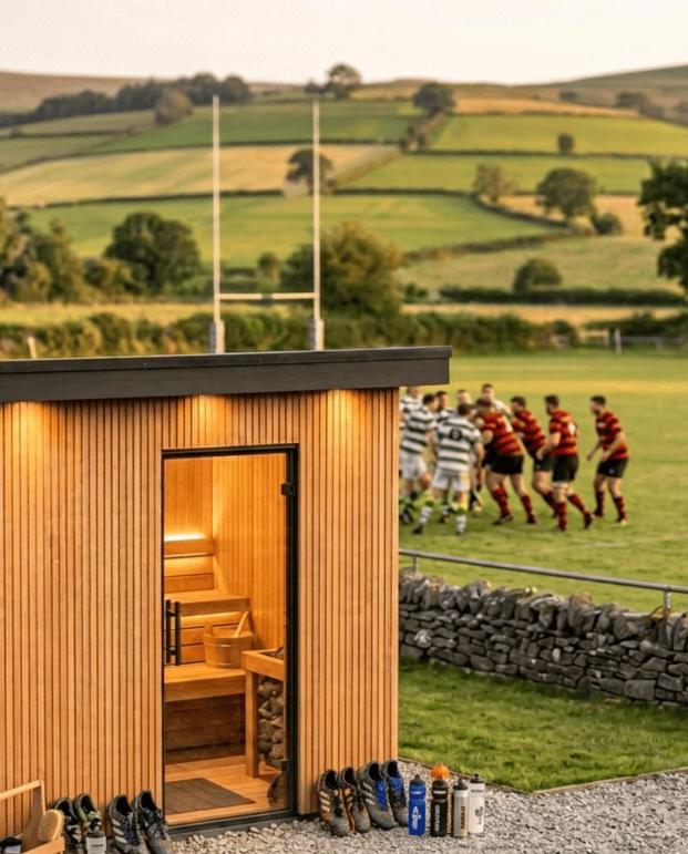 A small wooden building with an open door, revealing a sauna inside, situated on a grassy field. In the background, a rugby match is taking place with players in red and black striped uniforms and white and black striped uniforms, on a lush green field with rolling hills and trees.