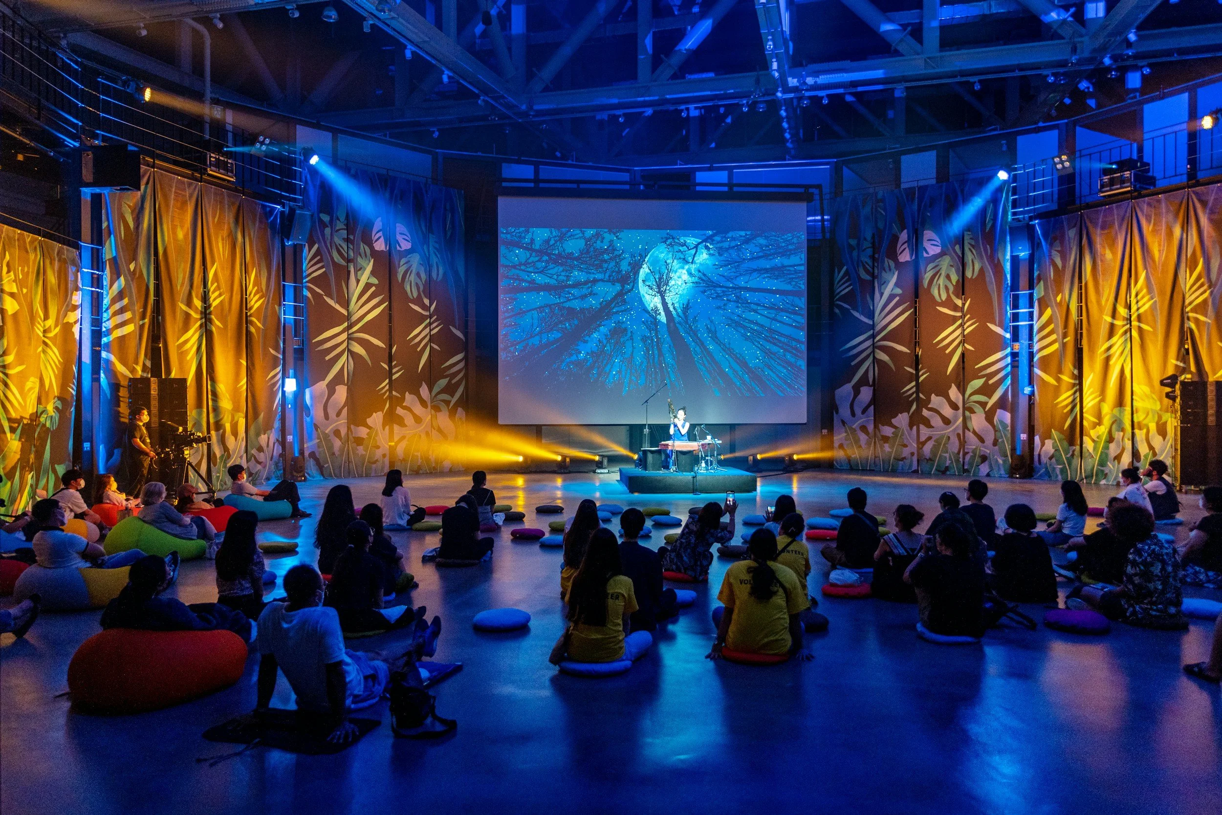 An indoor stage with a performer playing a traditional musical instrument, surrounded by colorful lighting and a large screen displaying a moon and tree scene. Audience members are seated on the floor with cushions, watching the performance.