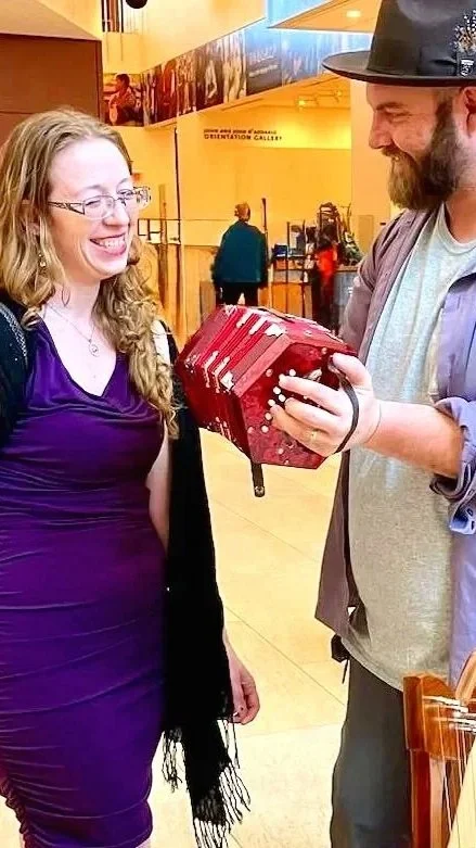 A young woman in a purple dress smiling as she looks at a man holding a red accordion with a vintage look, inside a brightly lit indoor venue.