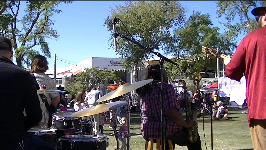 Musicians performing outdoors at a park during a festival, with a crowd of people and food stalls visible in the background.
