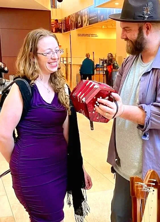 A man and a woman are smiling at each other inside a building. The man is holding a red accordion and is wearing a elegant hat, gray and purple shirt, and dress pants. The woman is wearing a purple dress with a black shawl and glasses.