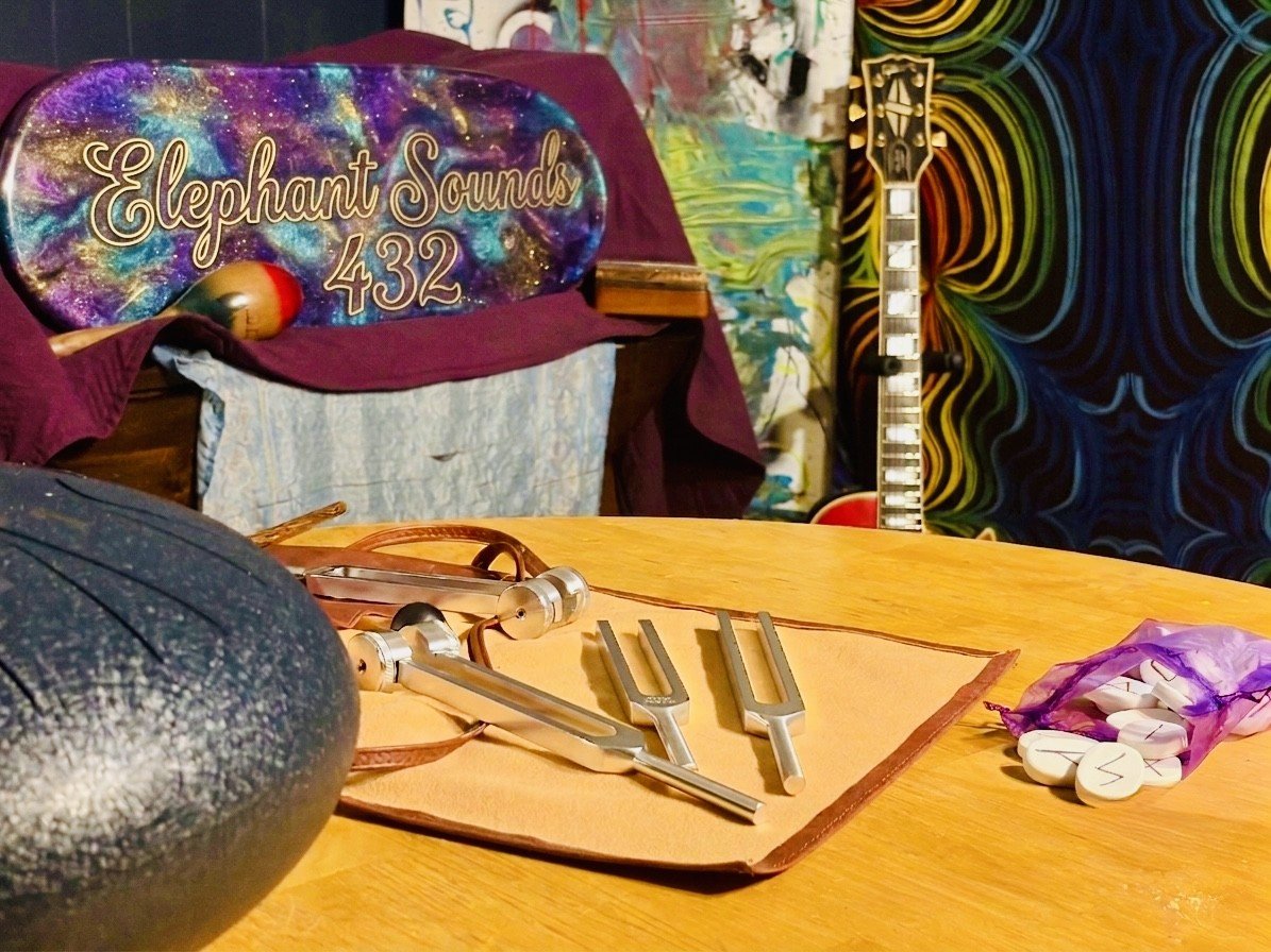 A wooden table with metal tongs, a game controller, and a purple drawstring bag filled with white pills labeled with a black lightning bolt. In the background, a colorful wall with a psychedelic pattern, a guitar, and a sign that reads "Elephant Sounds 432" on a display with a galaxy design.