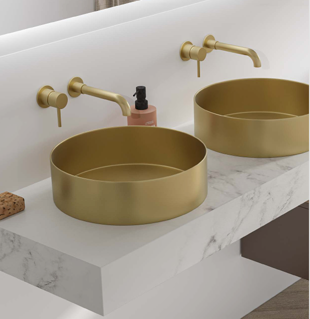 A modern bathroom double vanity with two round gold sinks, gold faucets, a pink soap dispenser, a sponge, and a marble countertop.