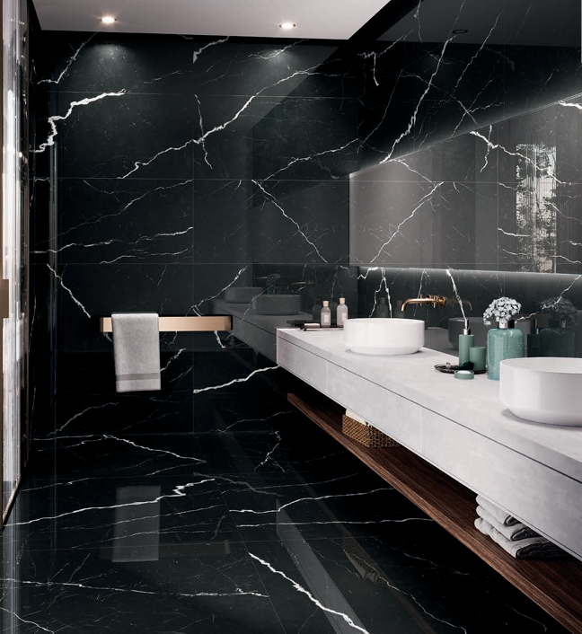Modern bathroom with black marble walls and floor, white countertop with two vessel sinks, wall-mounted gold faucet, large mirror, and decorative vases with flowers.