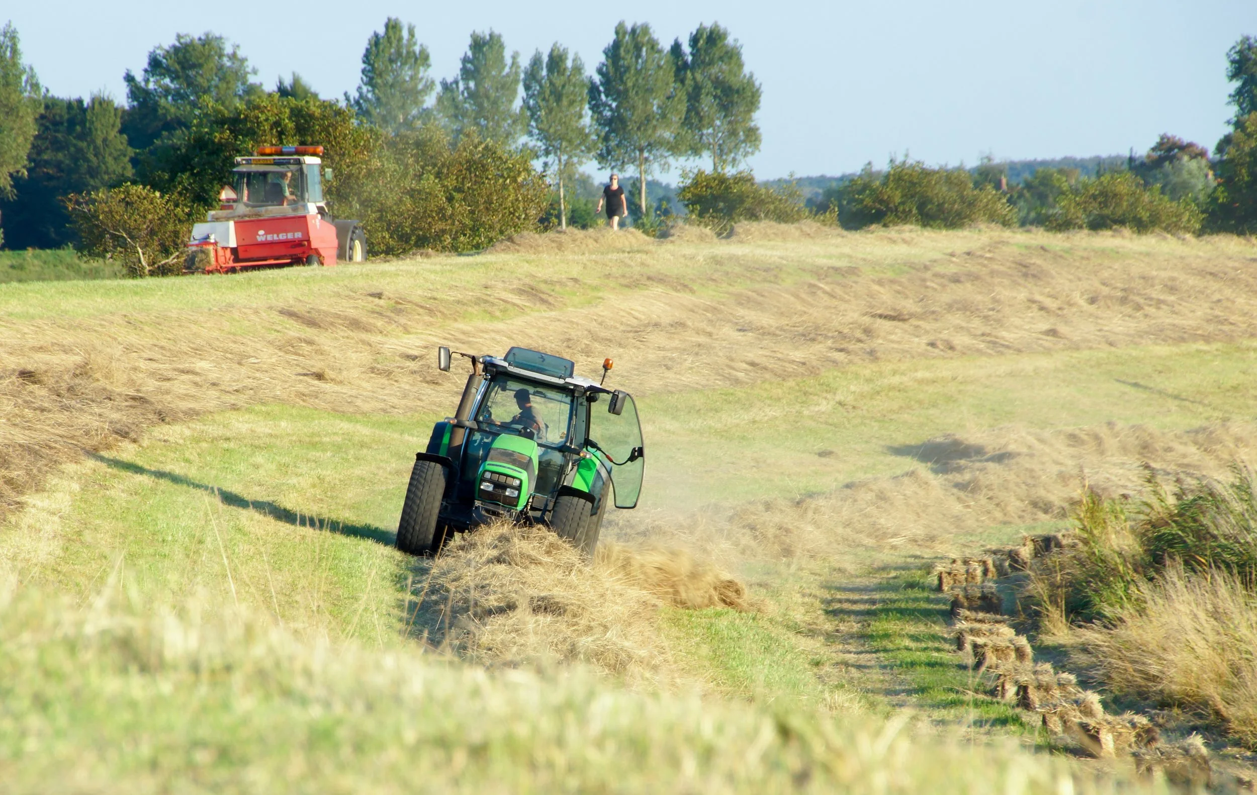 Twee tractors werken op een groen veld, waarbij gras wordt gehakseld of geoogst. Een persoon staat op de achtergrond.