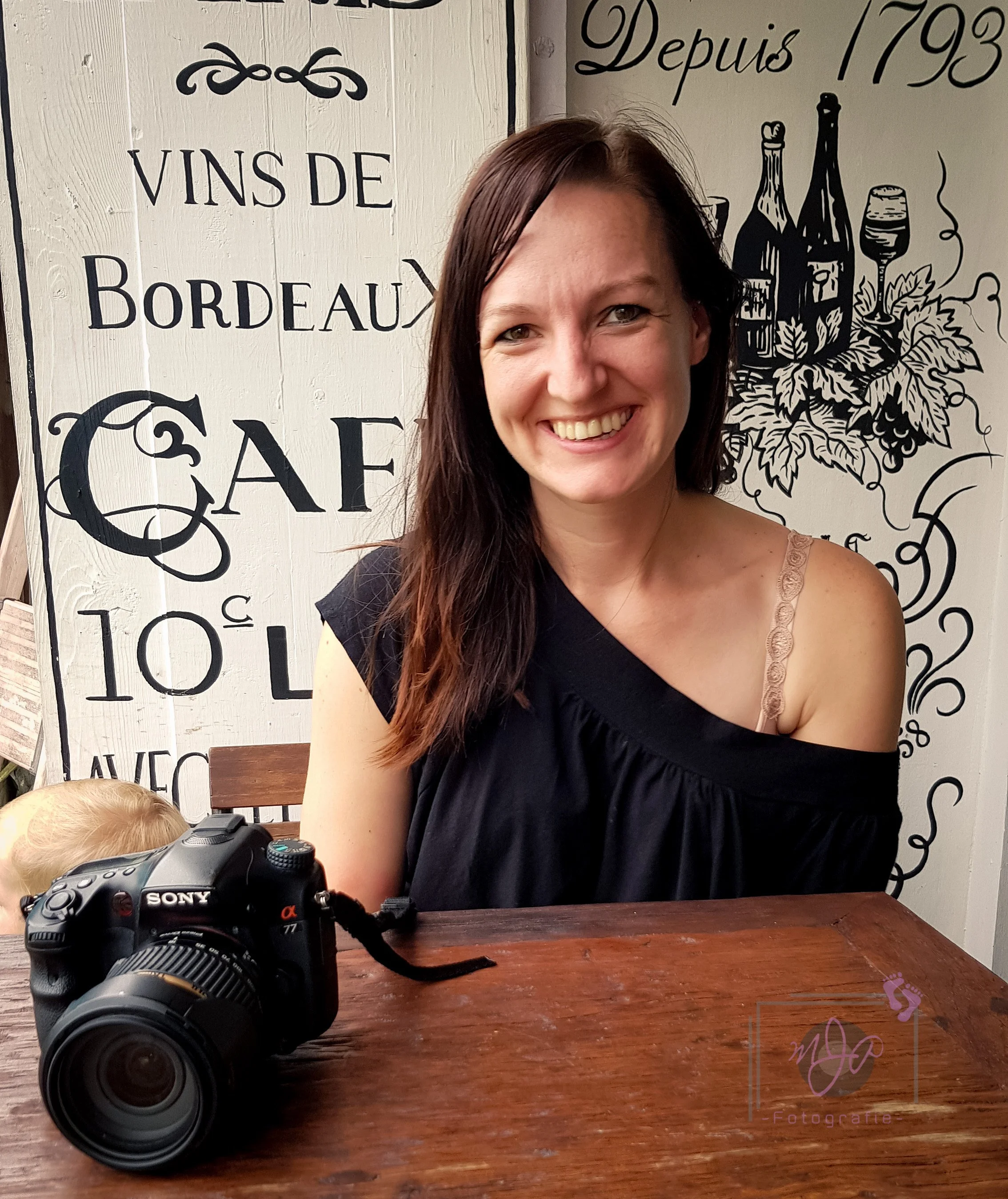 Een lachende vrouw met lang bruin haar zit aan een houten tafel, met een Sony-camera voor haar. Op de achtergrond een muur met Franse tekst en wijn-gerelateerde tekeningen.