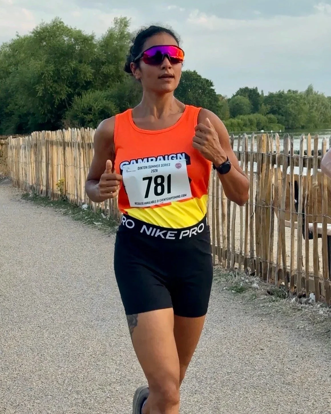 Female runner wearing sunglasses, an orange and yellow Nike Pro tank top, black shorts, and bib number 781, running along a gravel path with a wooden fence and green trees in the background.