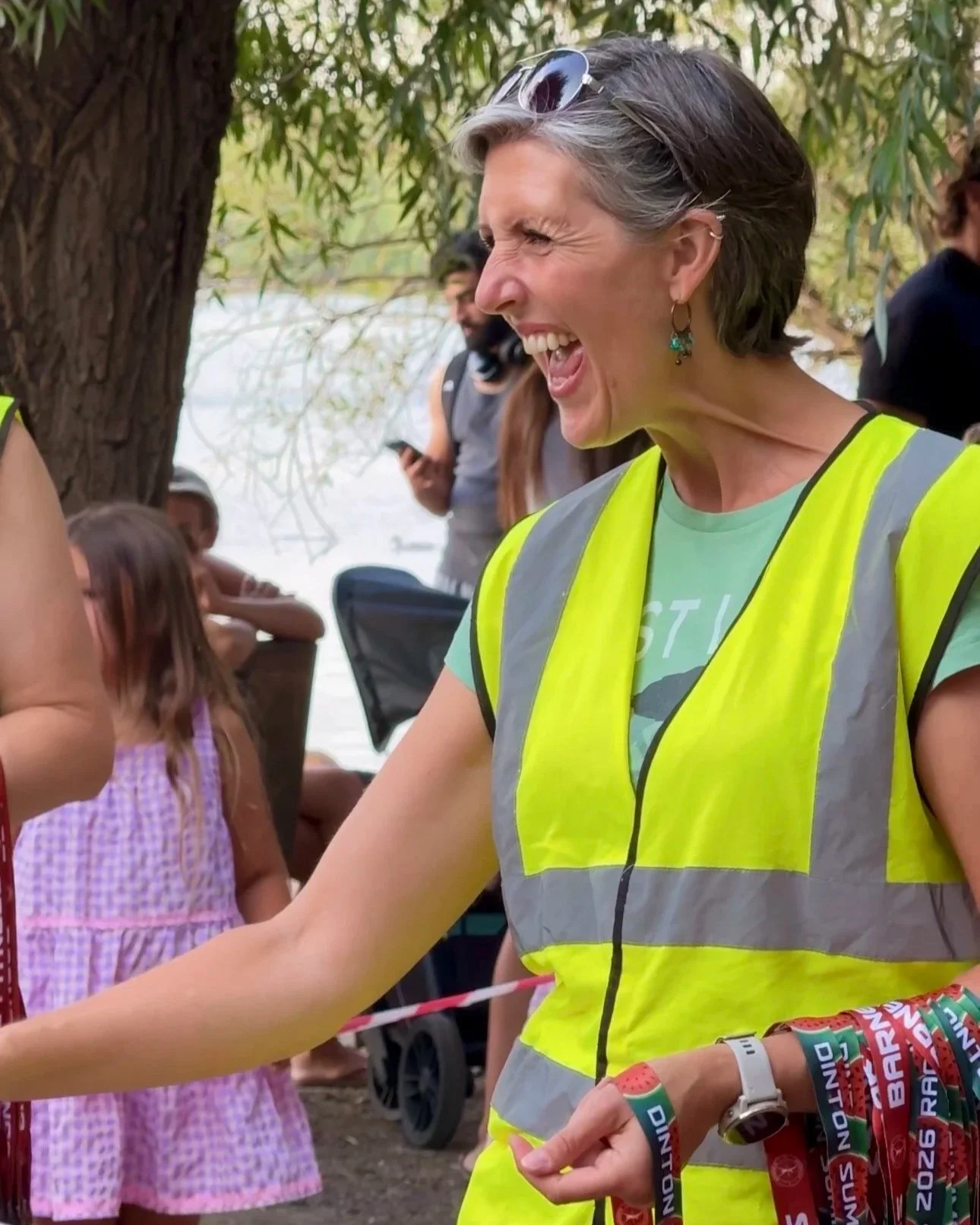 A woman with short gray hair, wearing sunglasses on her head and a yellow reflective safety vest, smiling and engaging in conversation outdoors near a tree, with children and other adults in the background.
