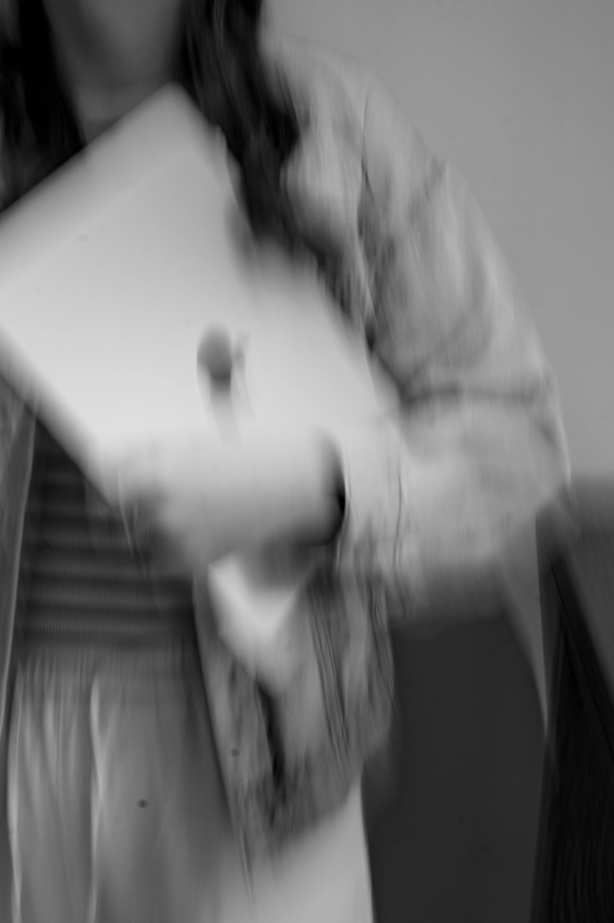 Blurry black-and-white photo of a person in a striped shirt and a jacket, with a hand resting on their cheek, sitting at a desk or table with a paper and pen.