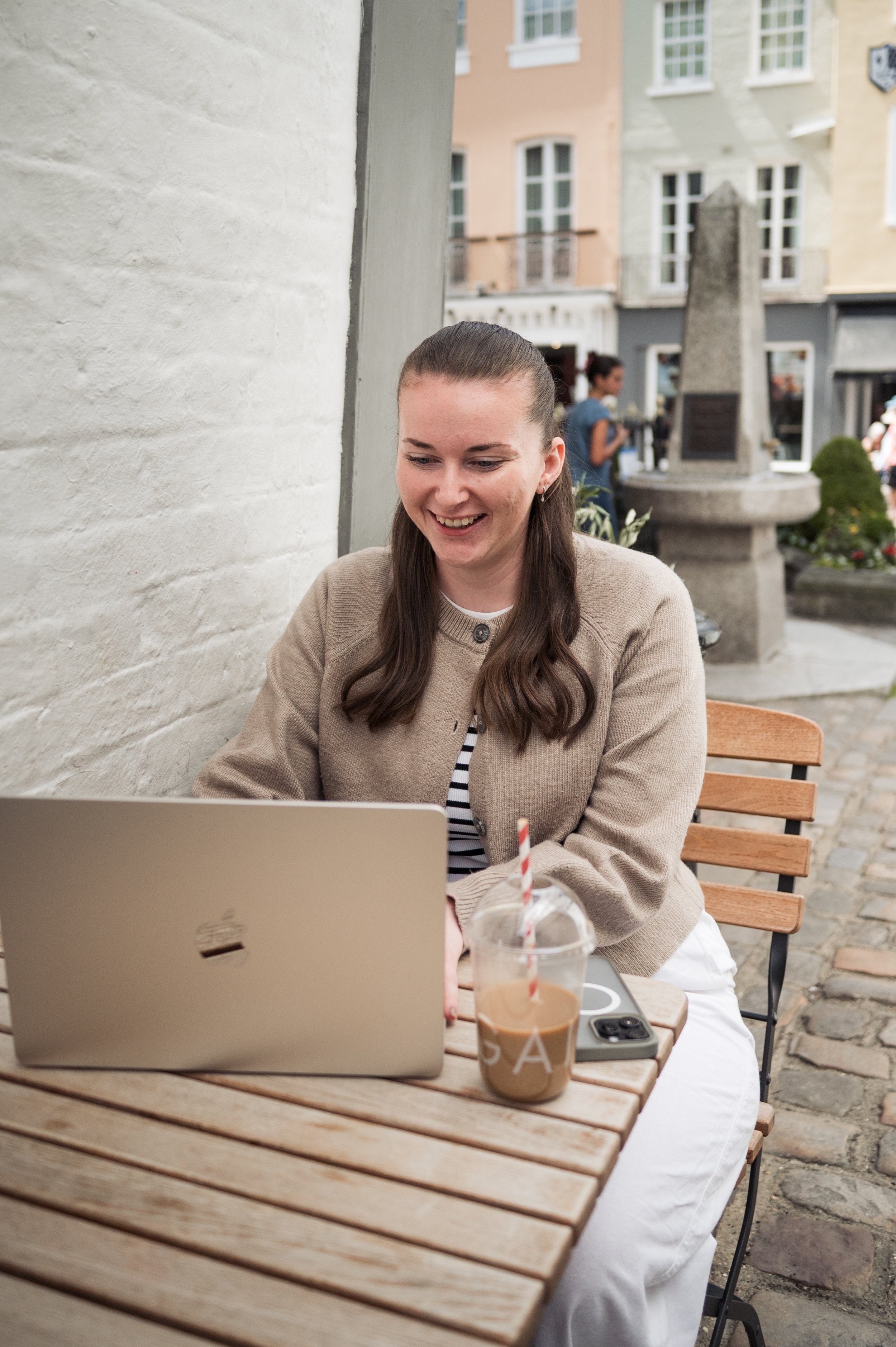 A woman with brown hair wearing a beige sweater and white pants is sitting at an outdoor table, smiling as she looks at her laptop. There is a coffee drink with a straw and a smartphone on the table in front of her. The background shows buildings with windows and a stone monument or fountain.