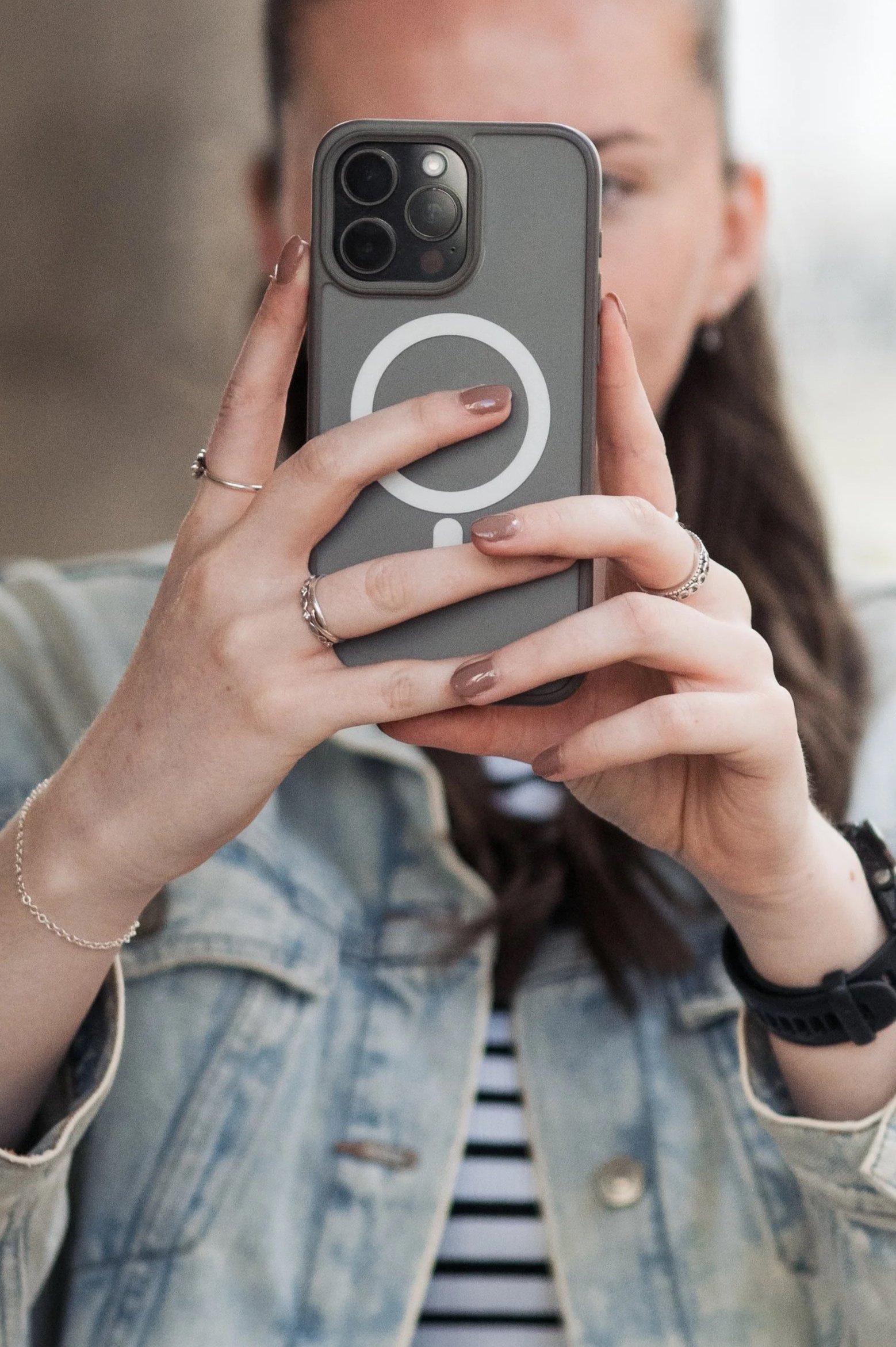 Person taking a mirror selfie with a smartphone, wearing a denim jacket and a black watch, with jewelry on their left hand.