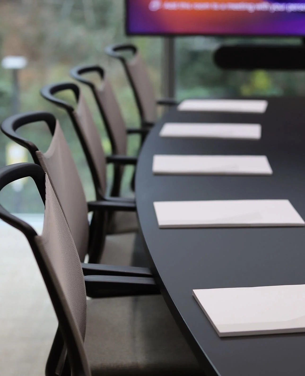 A conference room with a long dark table, beige chairs, and white papers evenly spaced on the table. A screen is visible in the background.