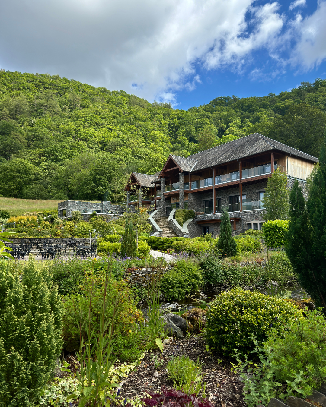 View of a large house with multiple balconies on a lush green hillside surrounded by trees and landscaping gardens with various plants and shrubs.