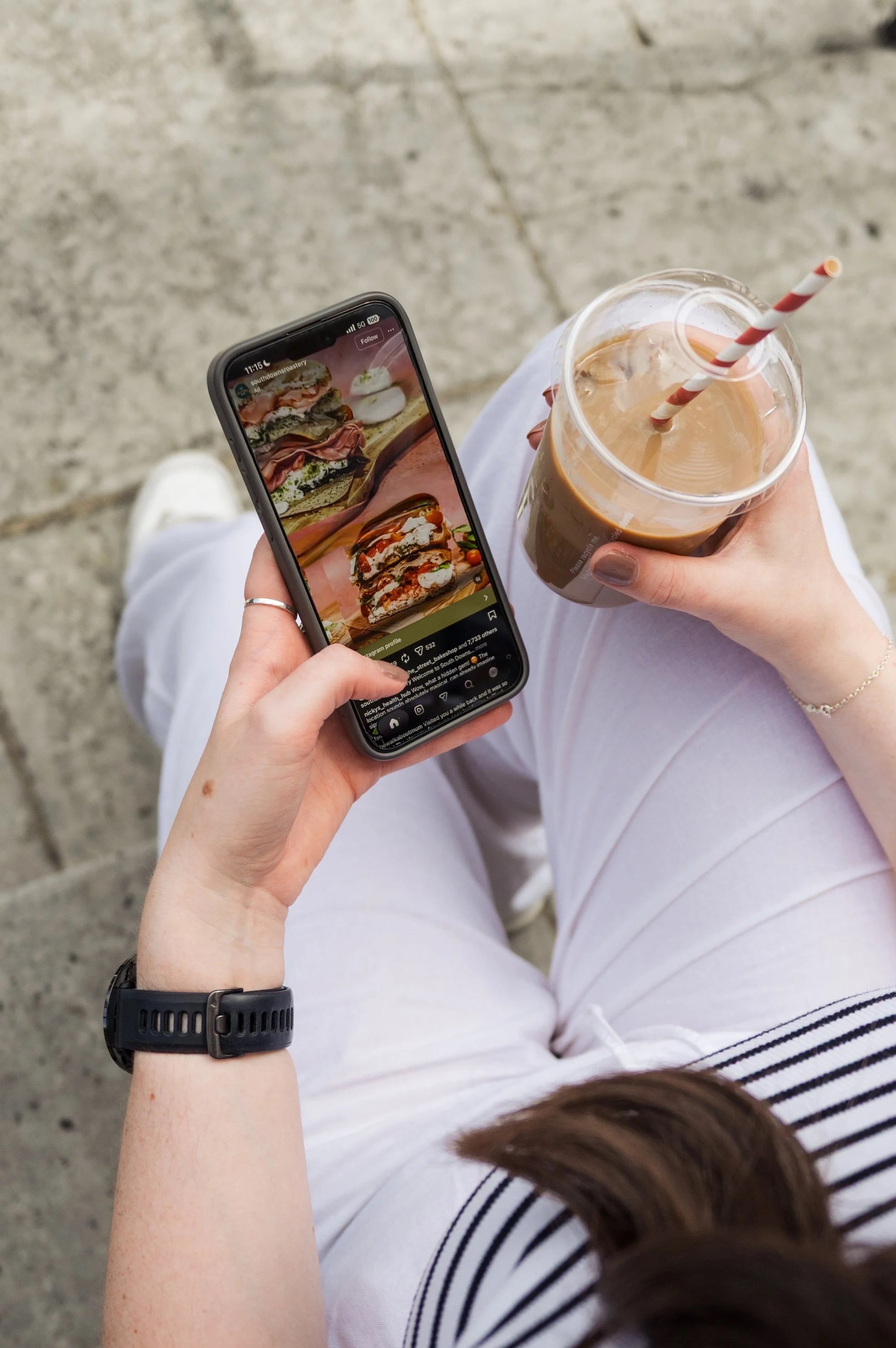 Person holding a smartphone displaying photos of sandwiches and a drink, sitting outdoors on a concrete surface, wearing white pants, a striped shirt, a black wristwatch, and holding a iced coffee beverage with a striped straw.