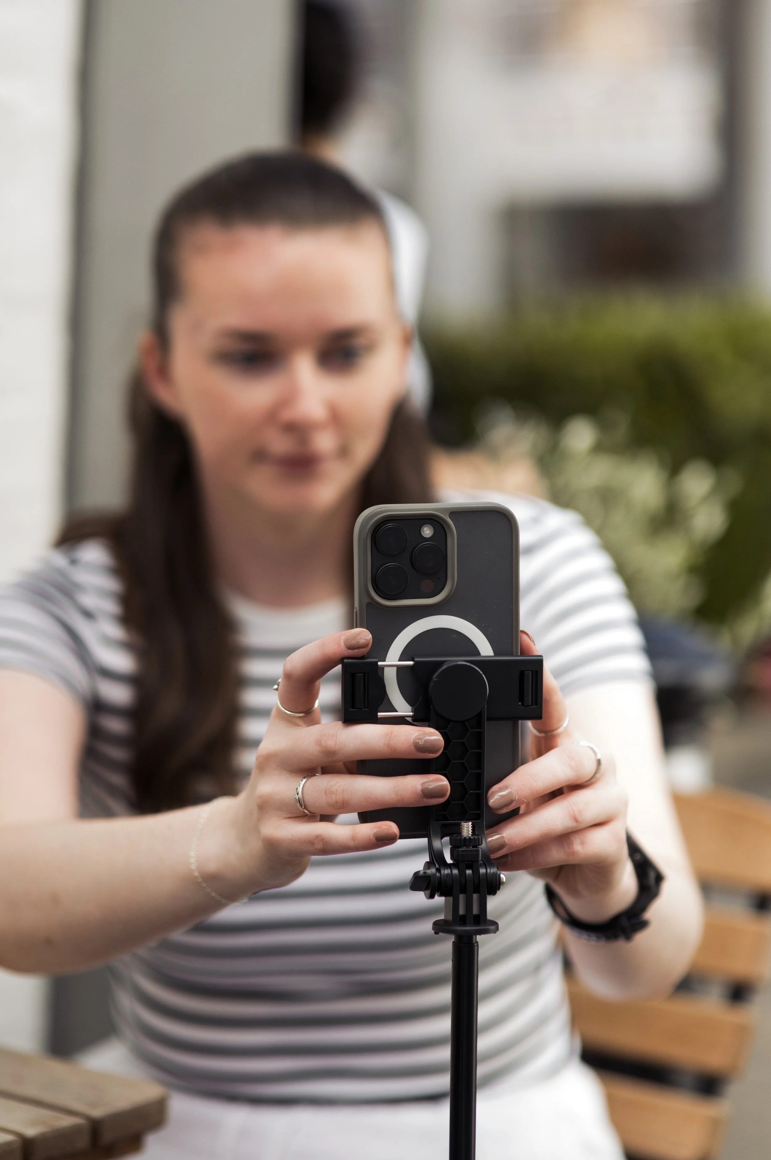 A woman in a striped shirt taking a selfie with her smartphone mounted on a tripod in an indoor setting.