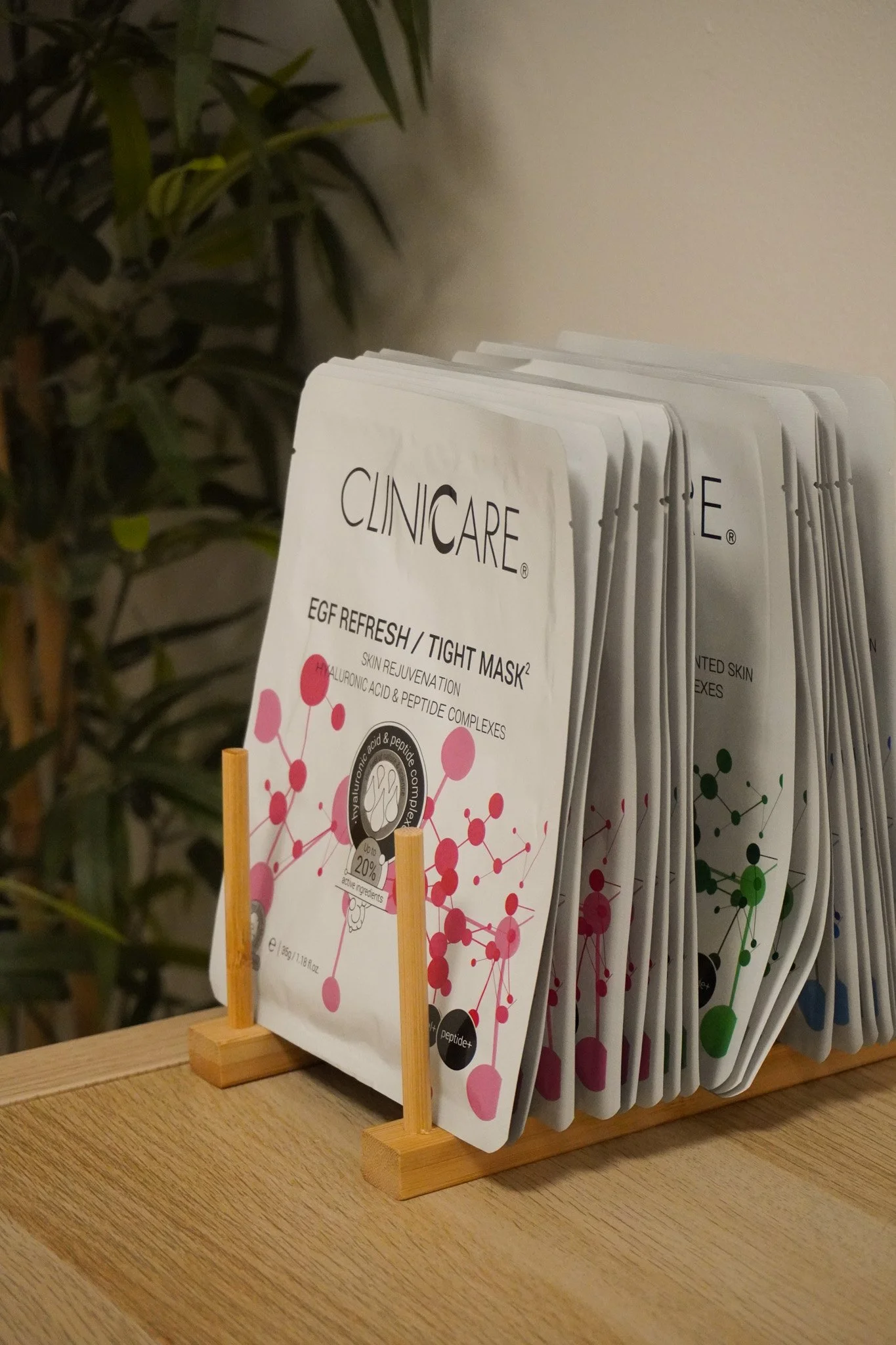 A display rack holding multiple packets of skincare masks, with the front packet labeled 'CINCARE' and 'EGF REFRESH / TIGHT MASK', placed on a wooden surface against a background of green leafy plants.