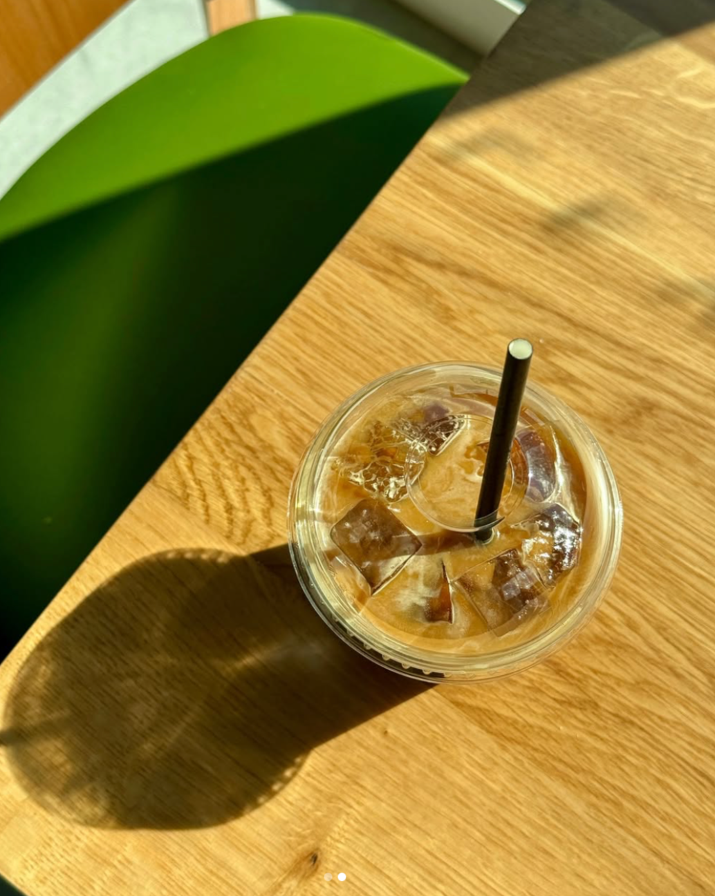 A cold iced coffee in a clear plastic cup with a black straw on a wooden table, casting a shadow.