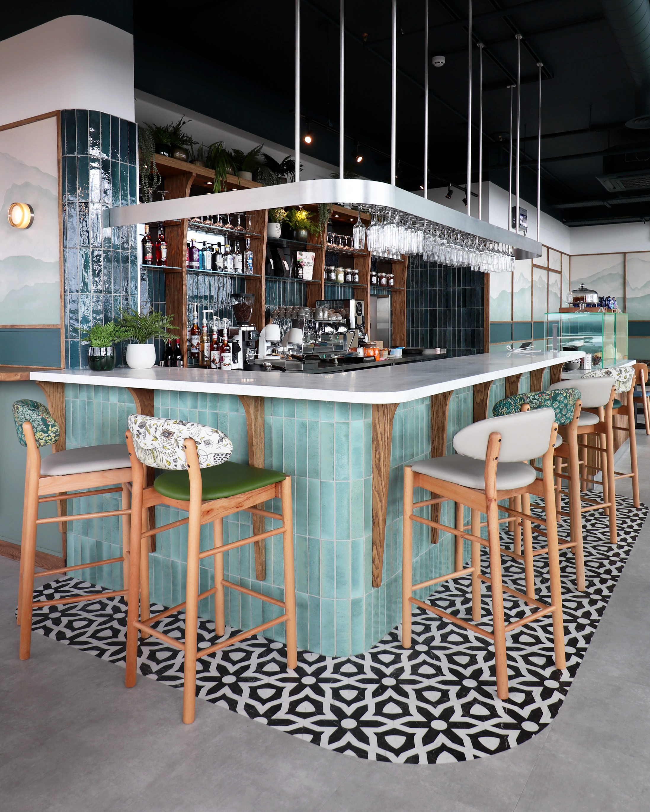 A modern bar interior with a white marble counter, teal tiled front, wooden accents, and black-and-white patterned flooring. High chairs with wooden frames and patterned fabric seats surround the bar, which has hanging glassware and potted plants on shelves behind it.