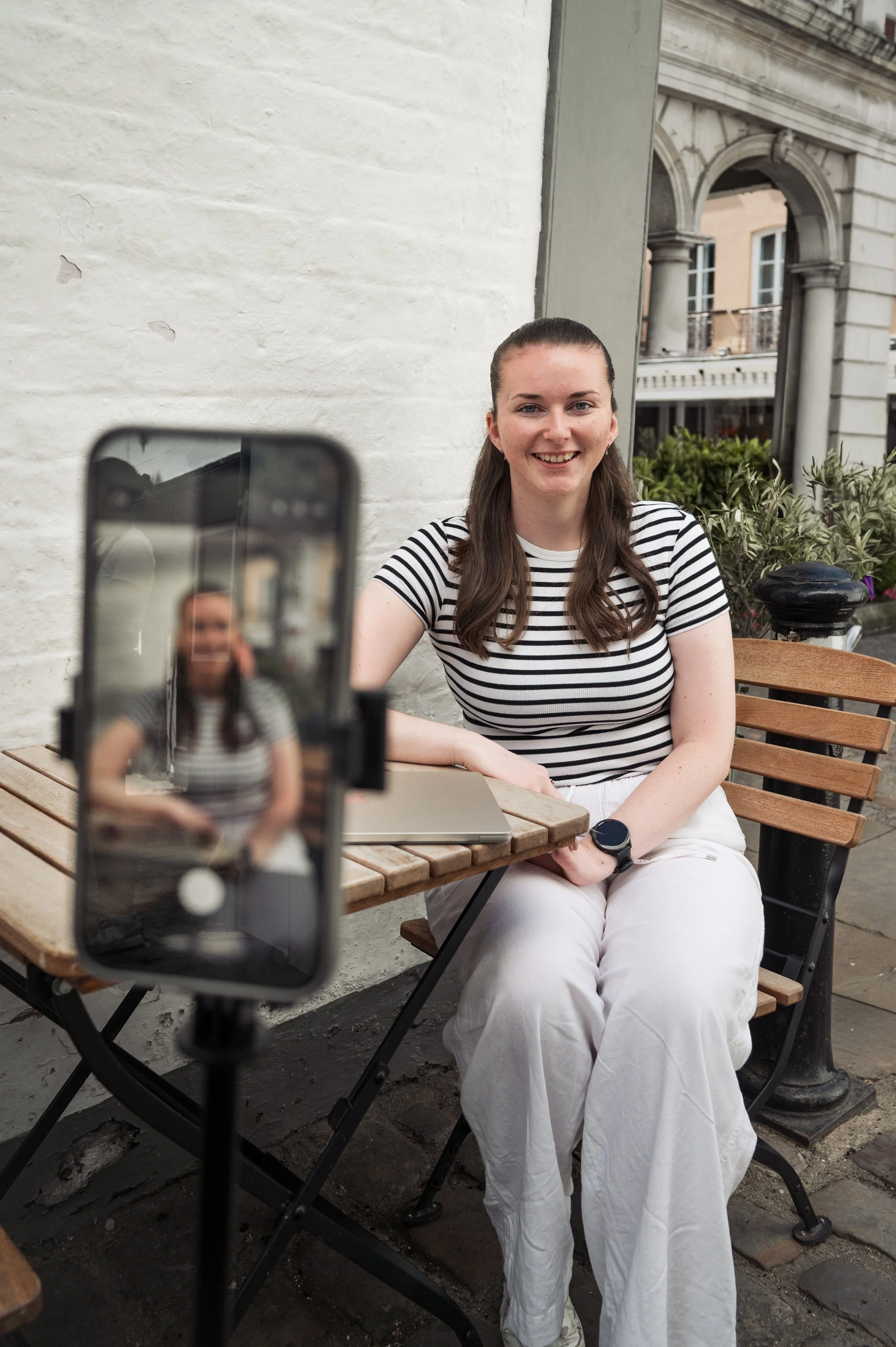A woman wearing a striped shirt and white pants sitting at an outdoor cafe table, smiling at the camera, with a smartphone on a tripod capturing her photo.
