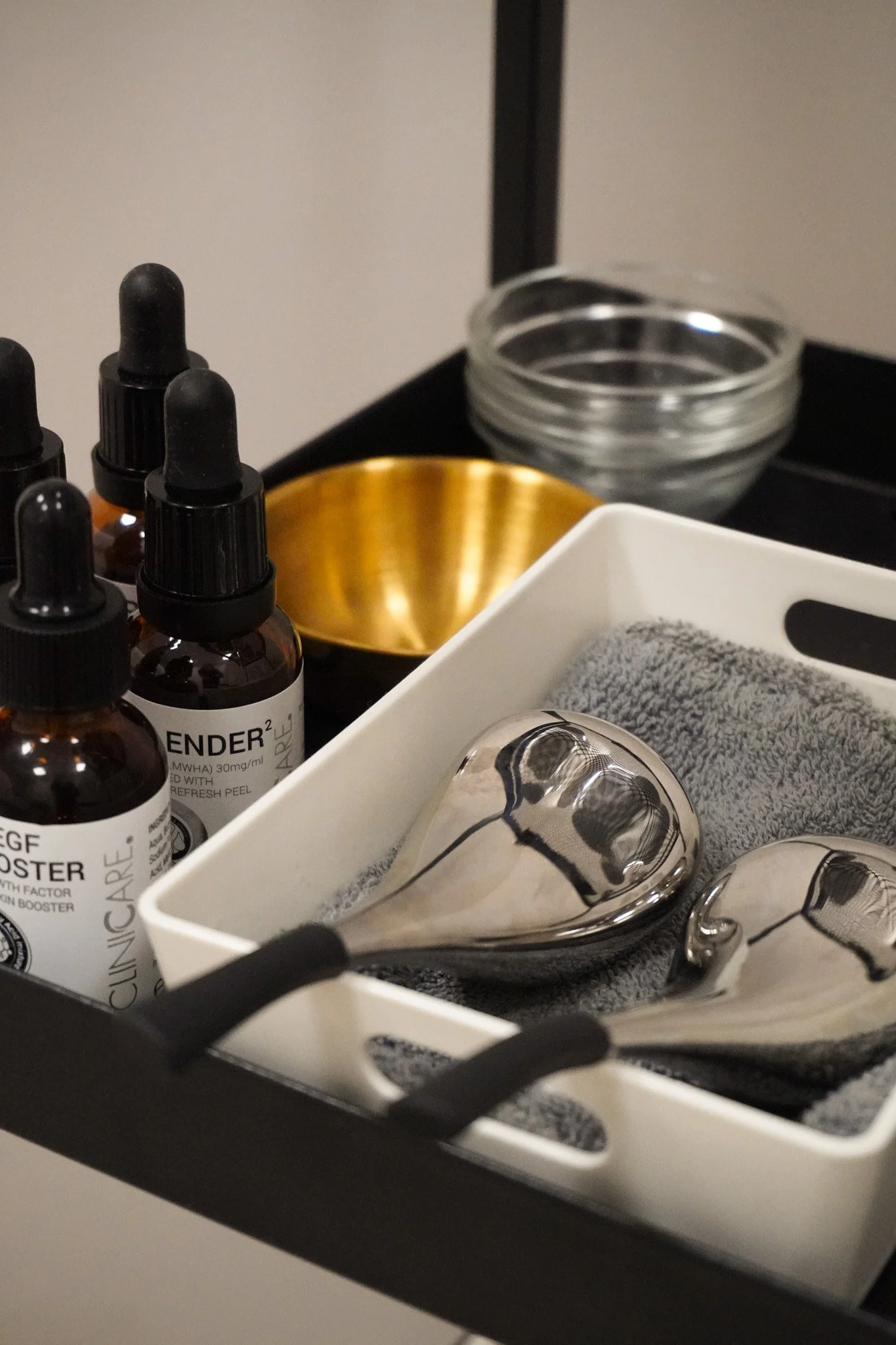 A collection of skincare or cosmetic bottles with droppers, a small stack of clear bowls, a white tray containing facial massage stones, and a gray towel, all arranged on a dark surface.