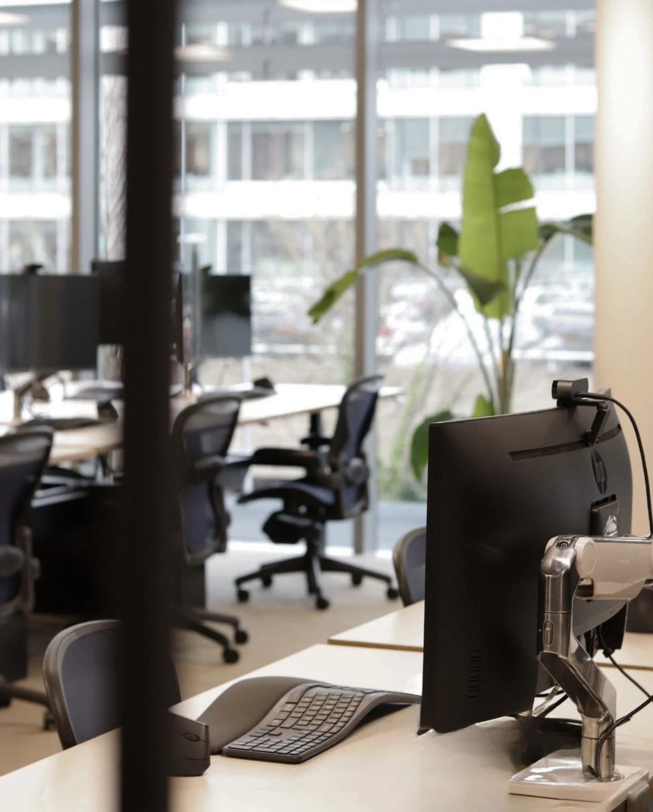 Office workspace with desks, chairs, computer monitors, and a large window with a view of a building and trees outside.