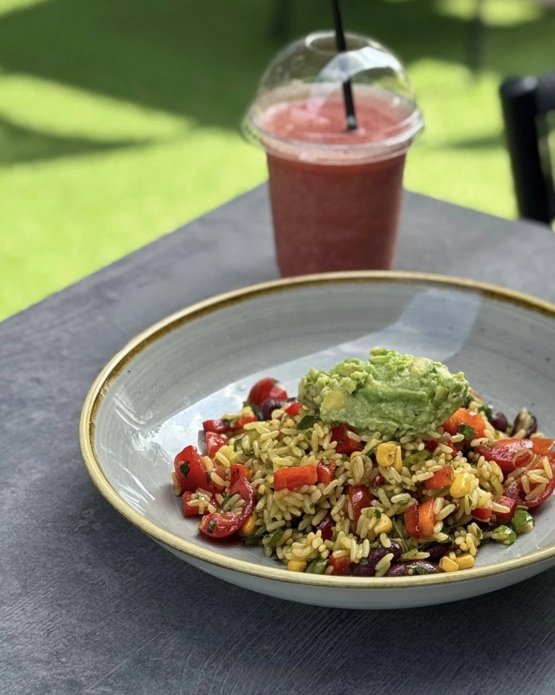 A plate of rice salad with chopped vegetables and a scoop of guacamole, with a pink fruit smoothie in a clear plastic cup with a lid and black straw, on a black table outdoors with green grass in the background.