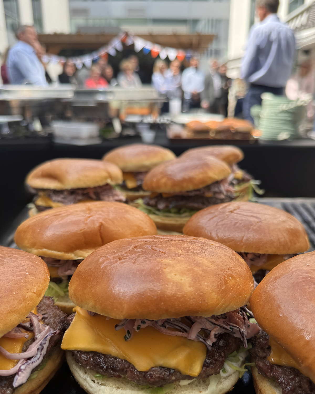 Close-up of several cheeseburgers with buns, beef patties, cheese, and shredded meat, on a grill at an outdoor event with people and a buffet in the background.