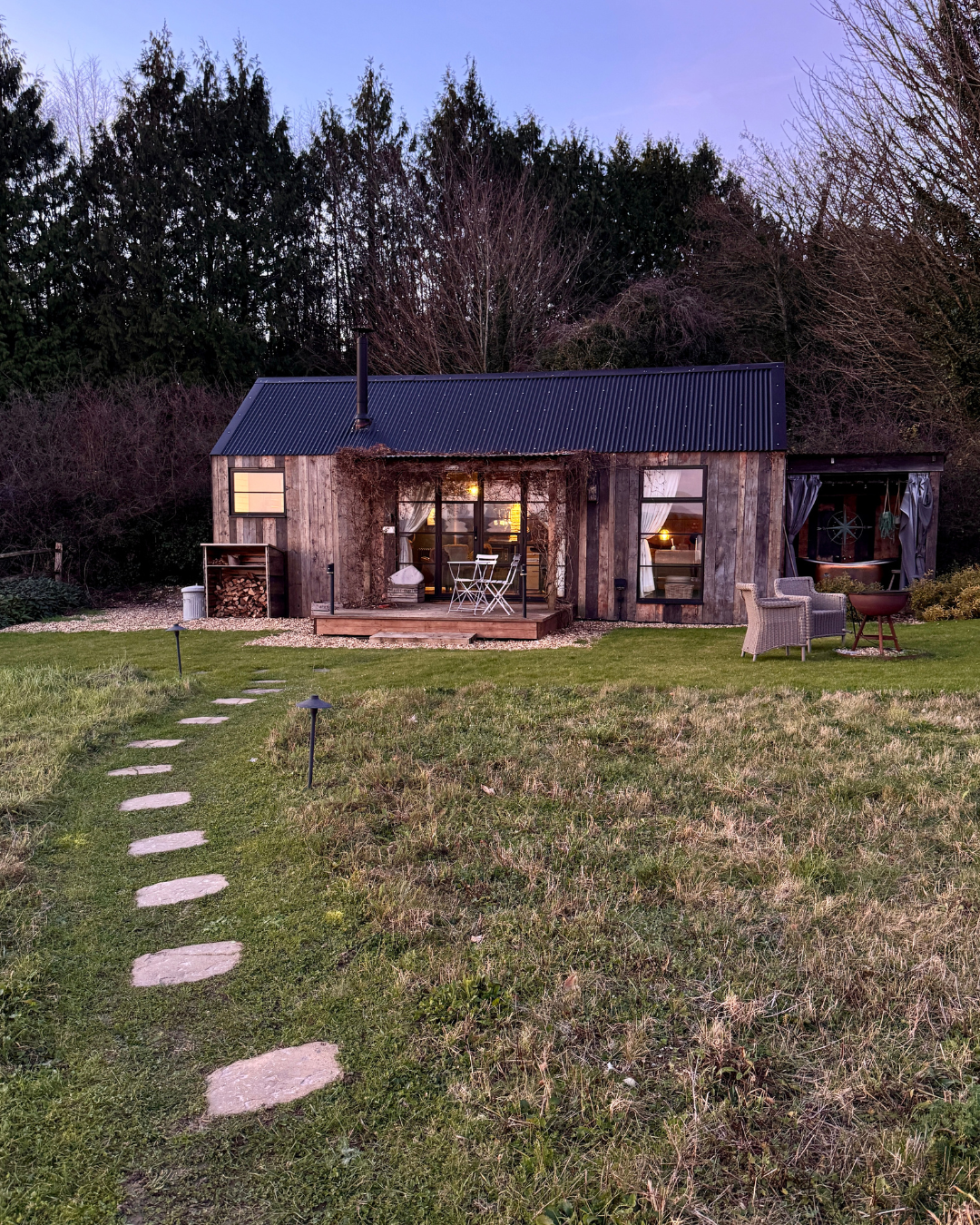 A rustic wooden house with a black metal roof, surrounded by a grassy yard with a stone pathway leading to the front porch. The house has large windows with white curtains, some lit from inside, and outdoor furniture including chairs and a fire pit. Trees are in the background during dusk.