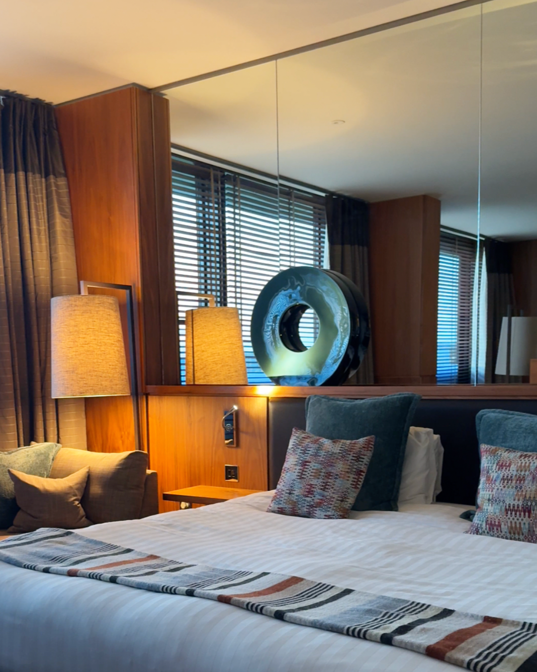 Hotel room with bed, pillows, bedside lamps, wooden wall paneling, and a decorative abstract sculpture reflected in a large mirror.
