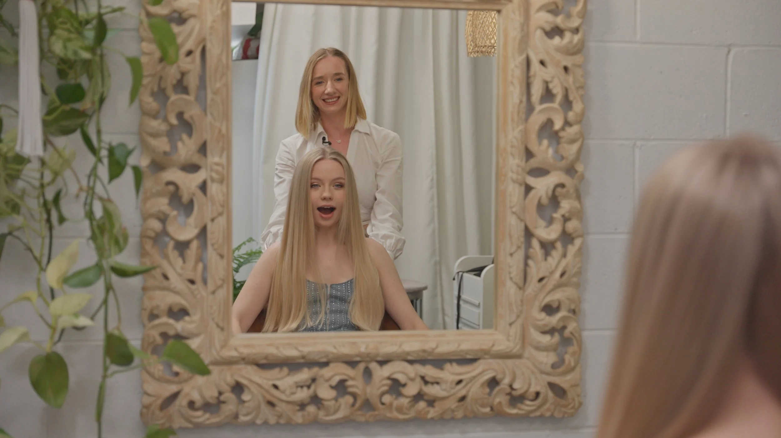 A woman with blonde hair sitting in front of a mirror while a hairstylist, standing behind her, styles her hair. The mirror has an ornate carved frame, and the background includes a white wall, curtains, and some green plants.