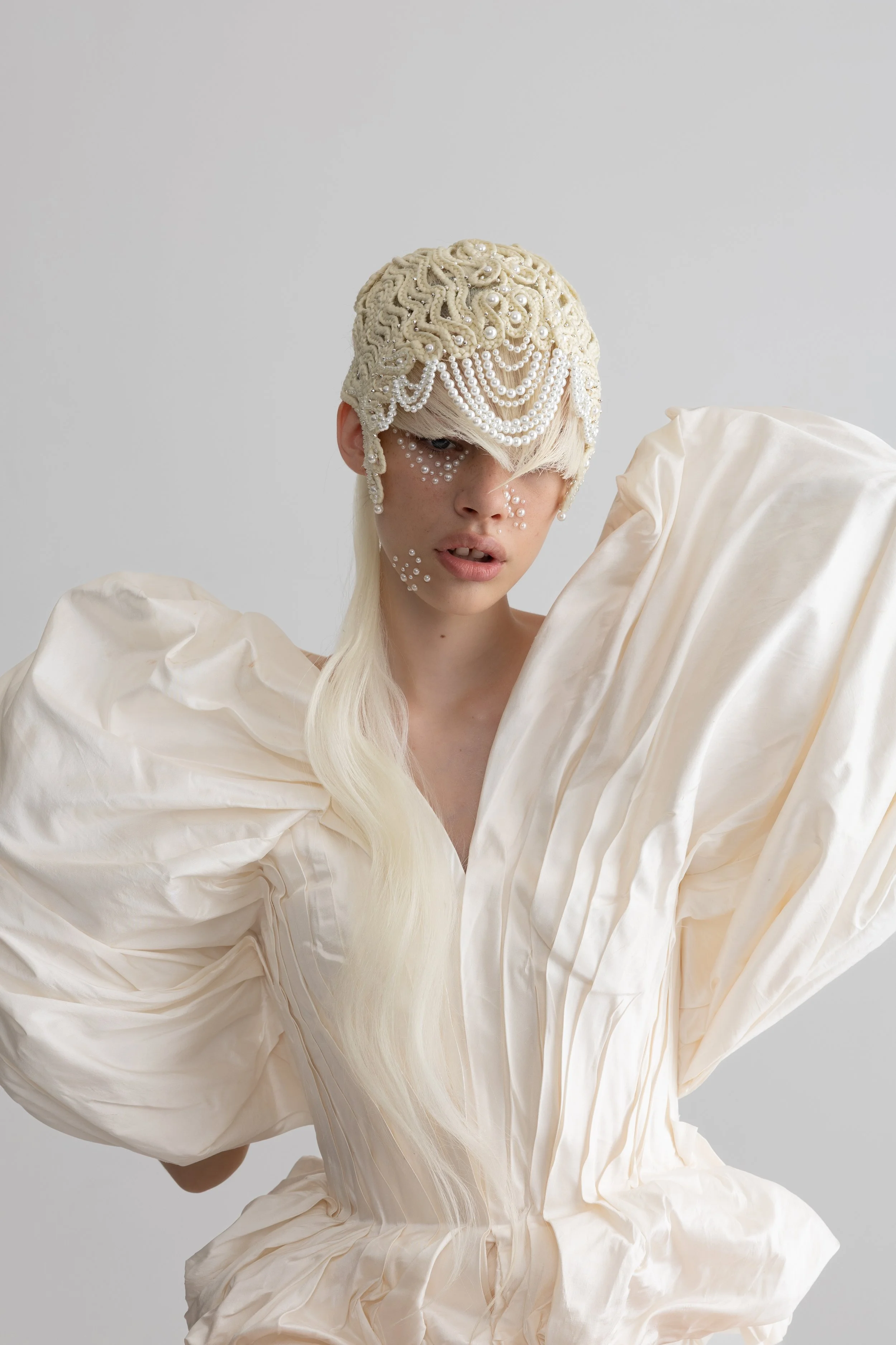A model wearing an elaborate cream-colored gown with large puffed sleeves and a designer headpiece decorated with pearls and intricate patterns, posed against a plain gray background.