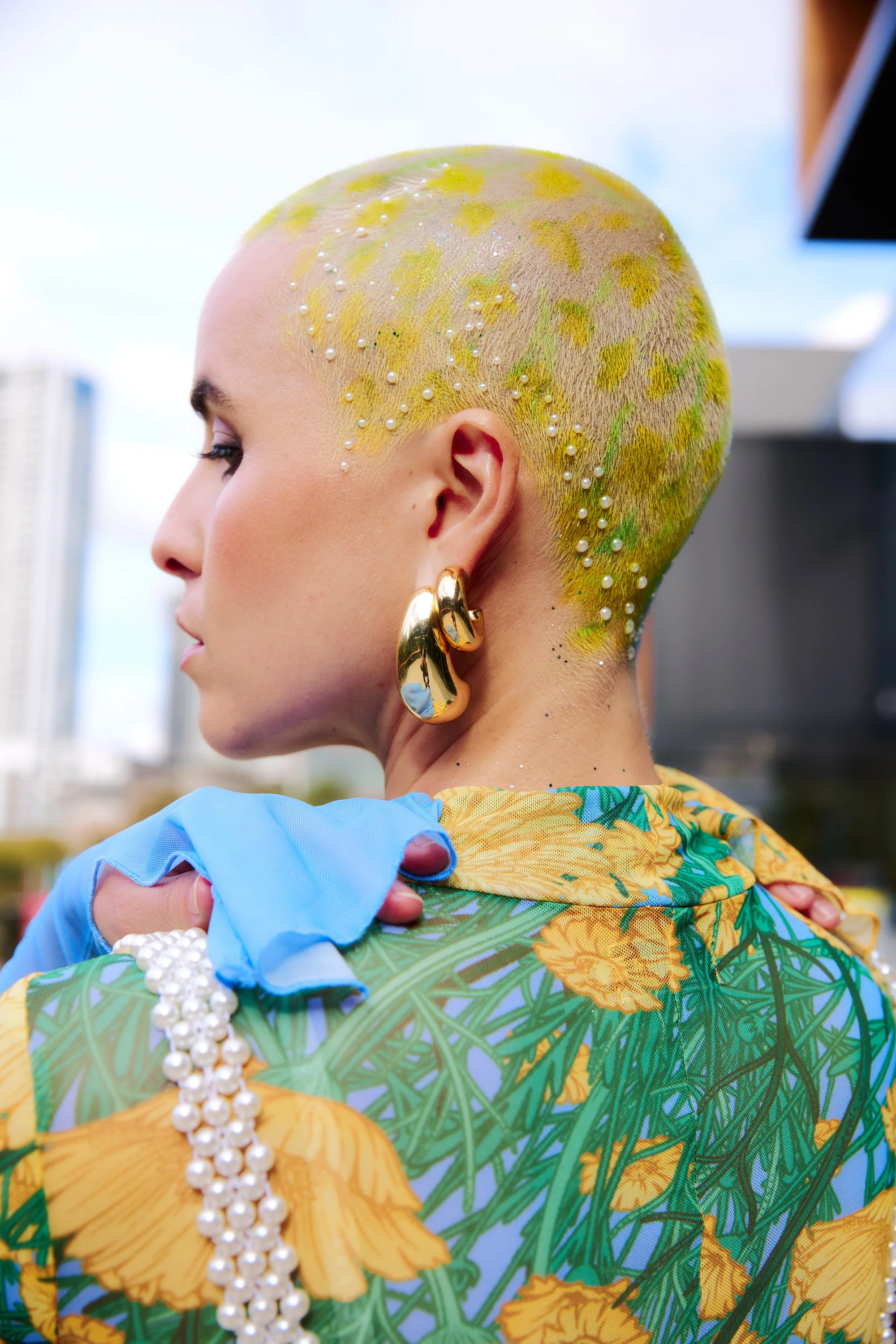 Profile view of a woman with a shaved head painted with yellow and green floral patterns, adorned with pearl embellishments, wearing gold earrings, and a colorful floral dress.