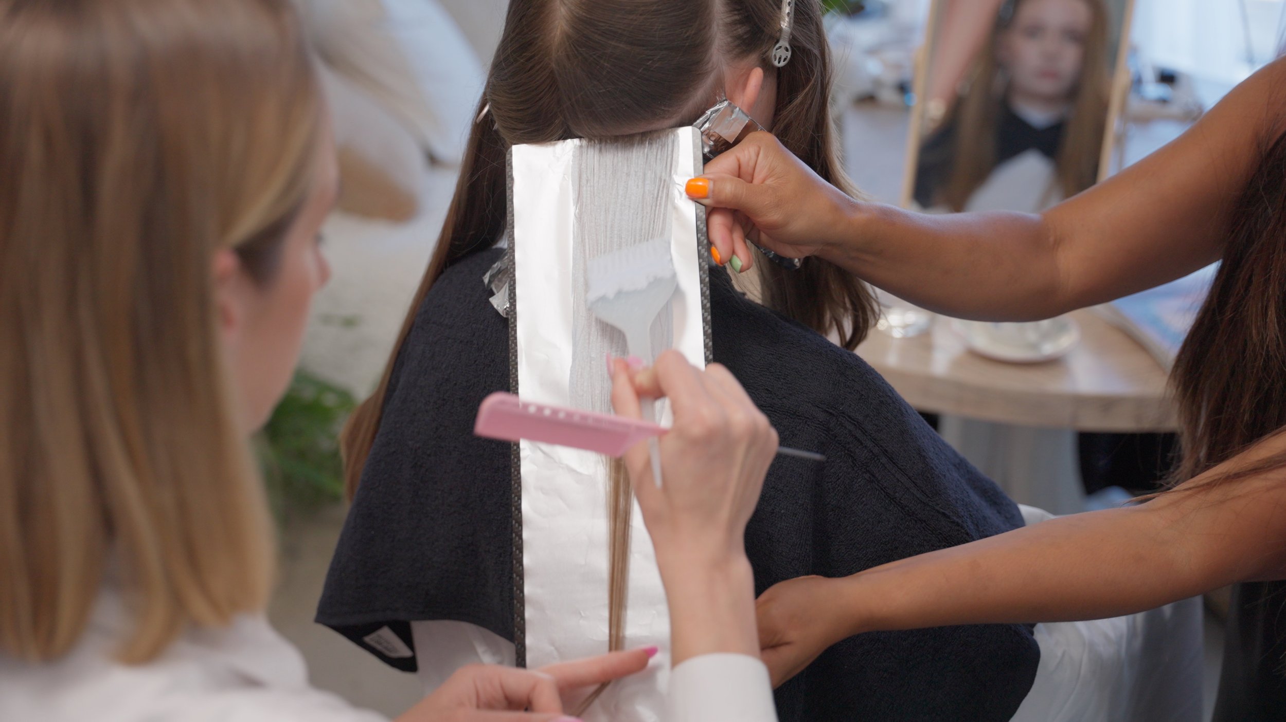 A person applying hair foil highlights to a woman's hair in a salon. The salon is busy, with other people and a mirror visible in the background.