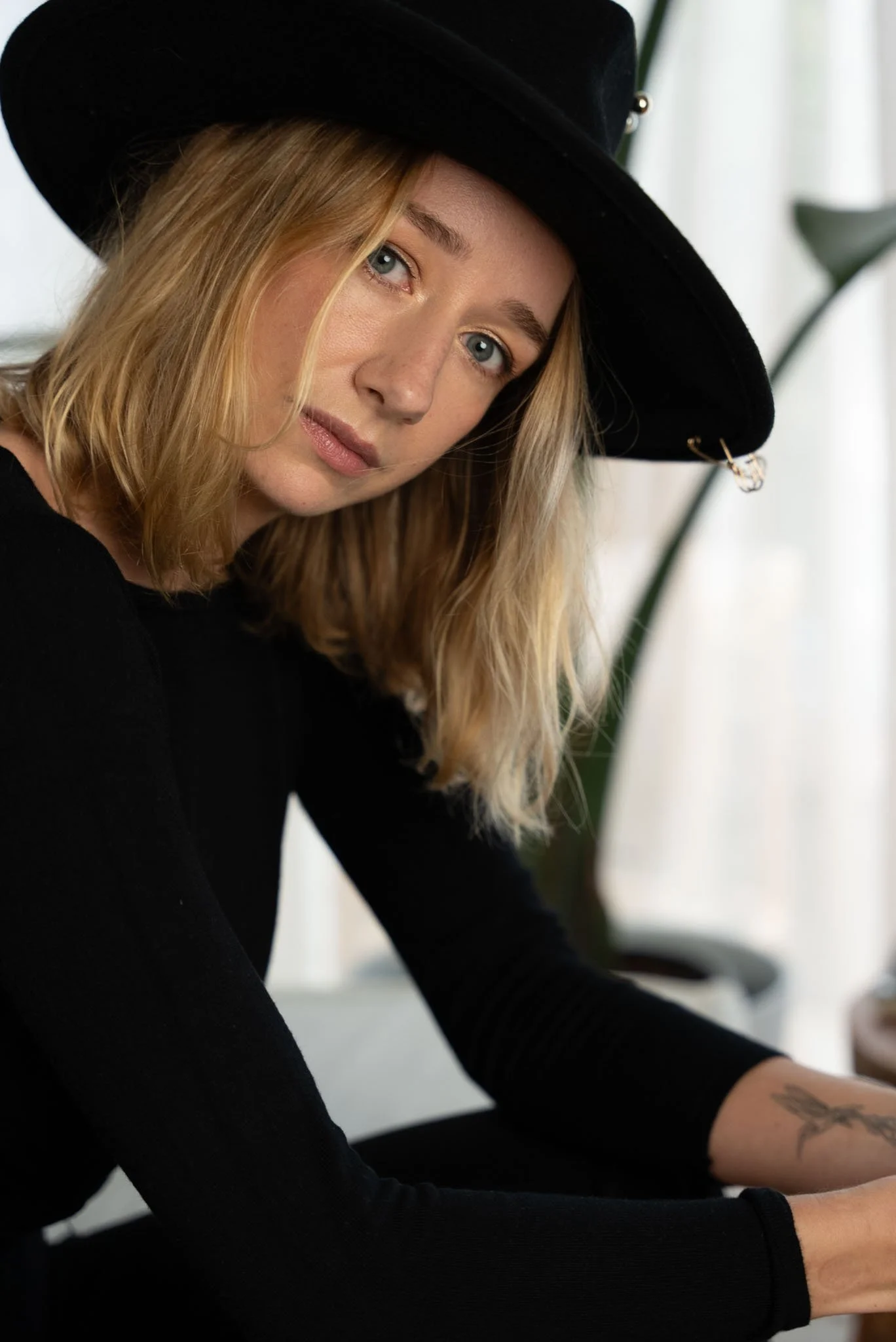 Close-up of a woman with blonde hair wearing a black hat and black long sleeve shirt, sitting at a desk with a tattoo visible on her left forearm, in a bright room with green plant leaves in the background.