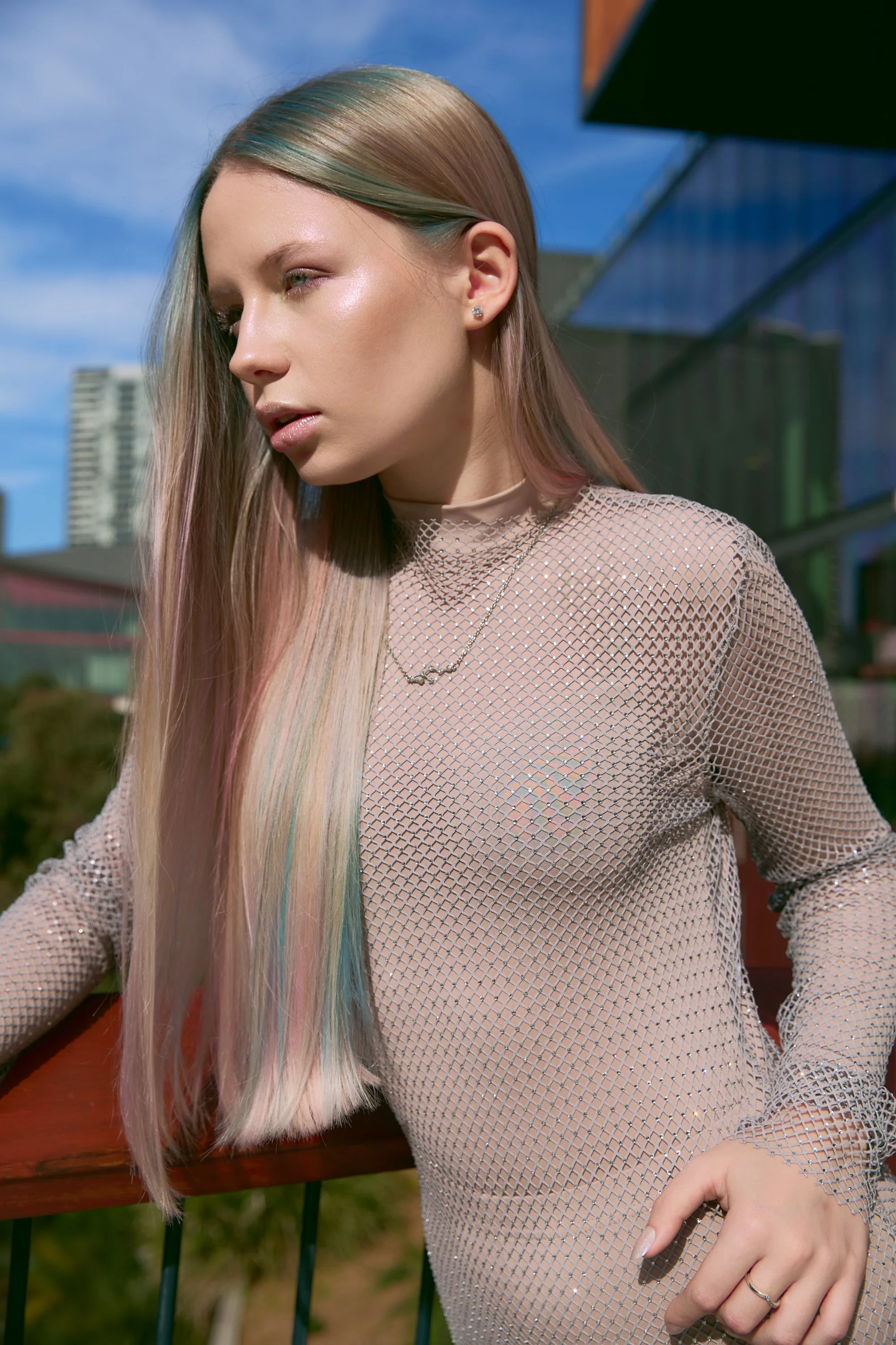 Close-up of a woman with long iridescent hair, wearing a see-through, sparkly top, standing outdoors with a building in the background.