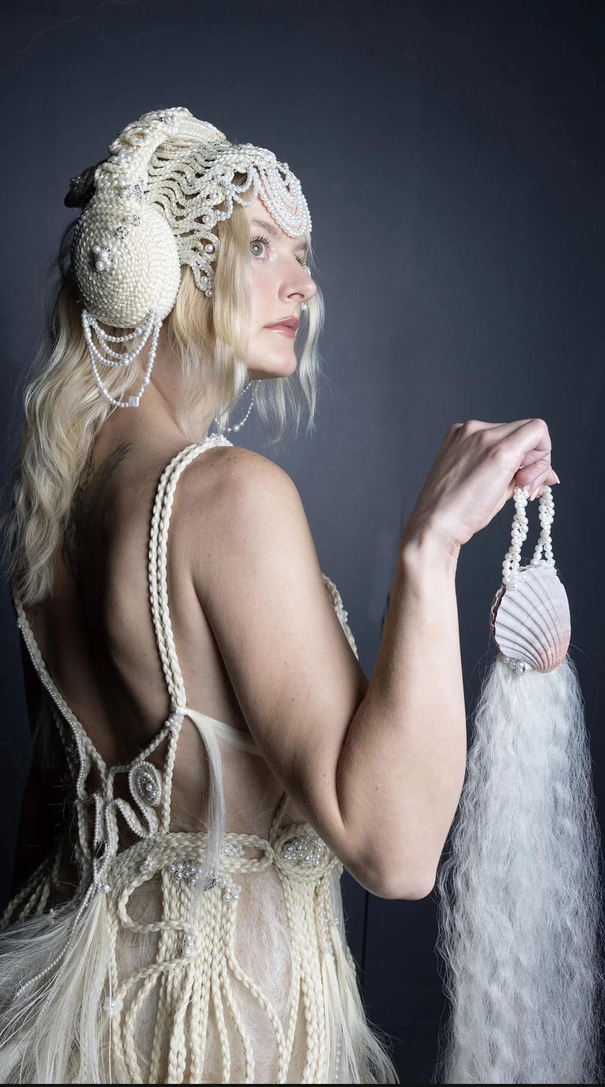 A woman with long blonde hair wearing an elaborate headdress and dress made of white braided and beaded material, holding a seashell purse with a long feathered tail.