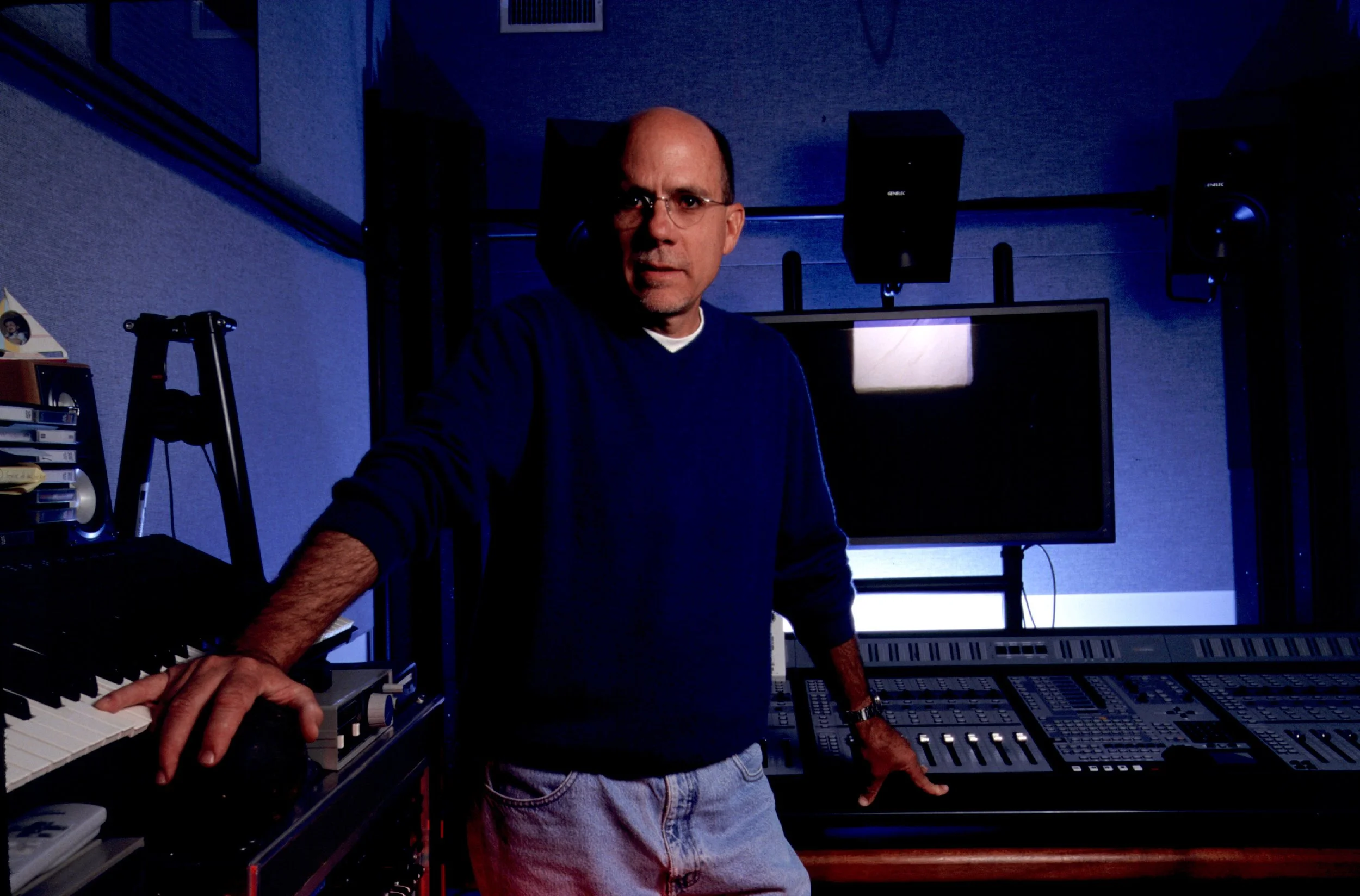 A man in glasses and a dark sweater standing in a music studio with audio equipment, speakers, a keyboard, and a computer monitor.