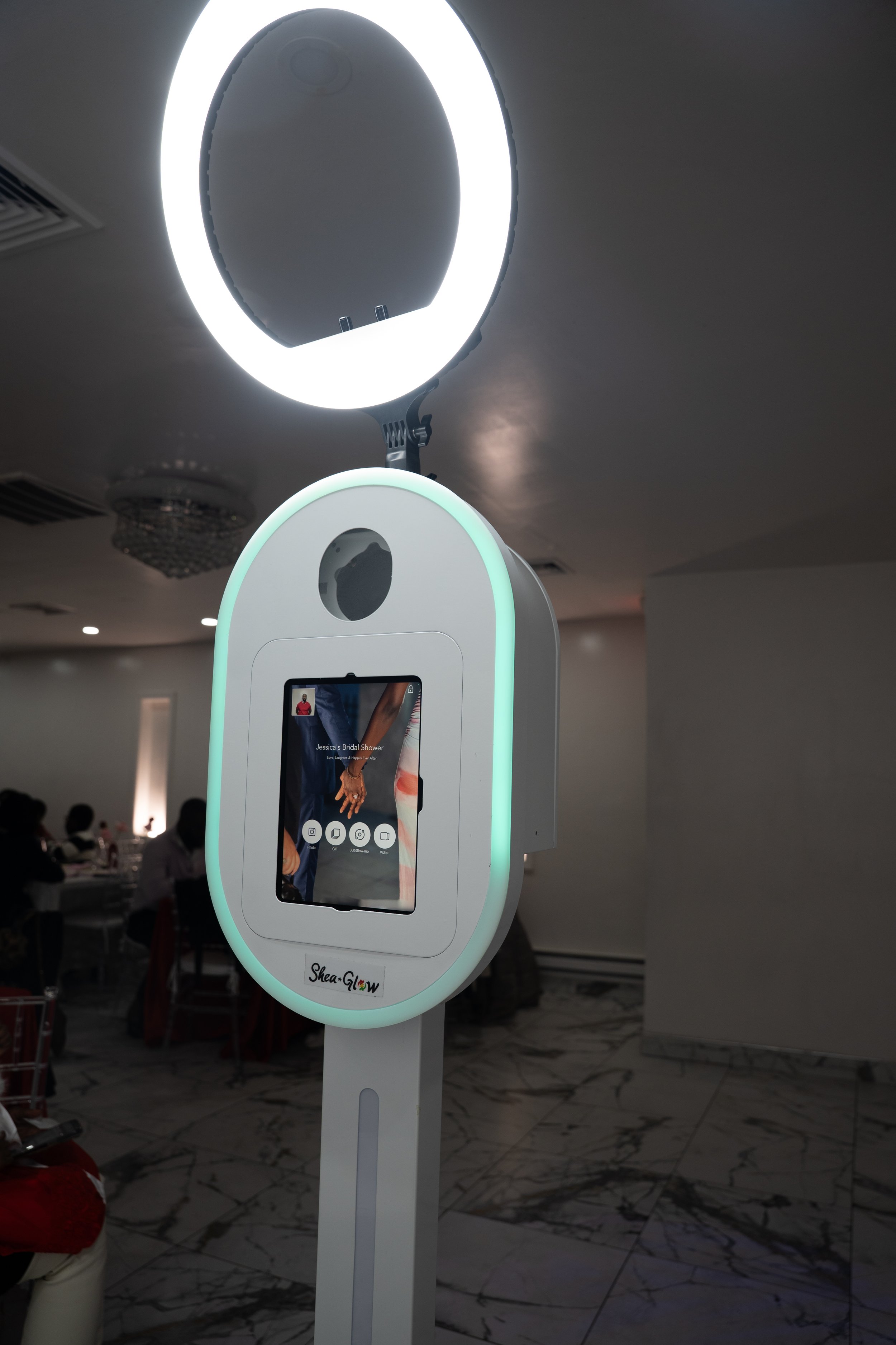 A digital photo booth with a circular ring light, located indoors, with people sitting at tables in the background.
