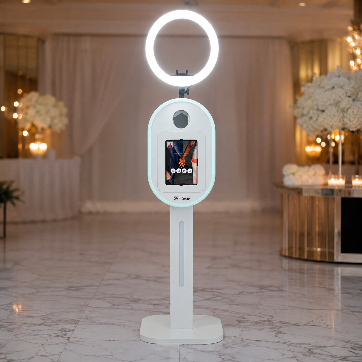 A white, modern COVID-19 temperature screening kiosk with a circular light at the top, a camera or sensor below it, and a touchscreen display in the middle, set in an elegant, well-lit indoor venue decorated with white flowers and candles.