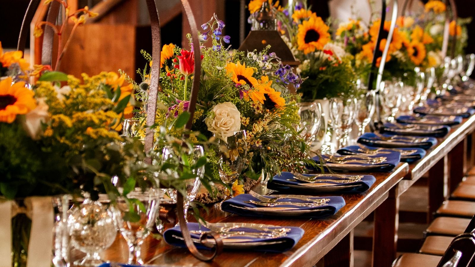 A long wooden dining table decorated with colorful flower arrangements and set with blue napkins and silverware for a formal event.