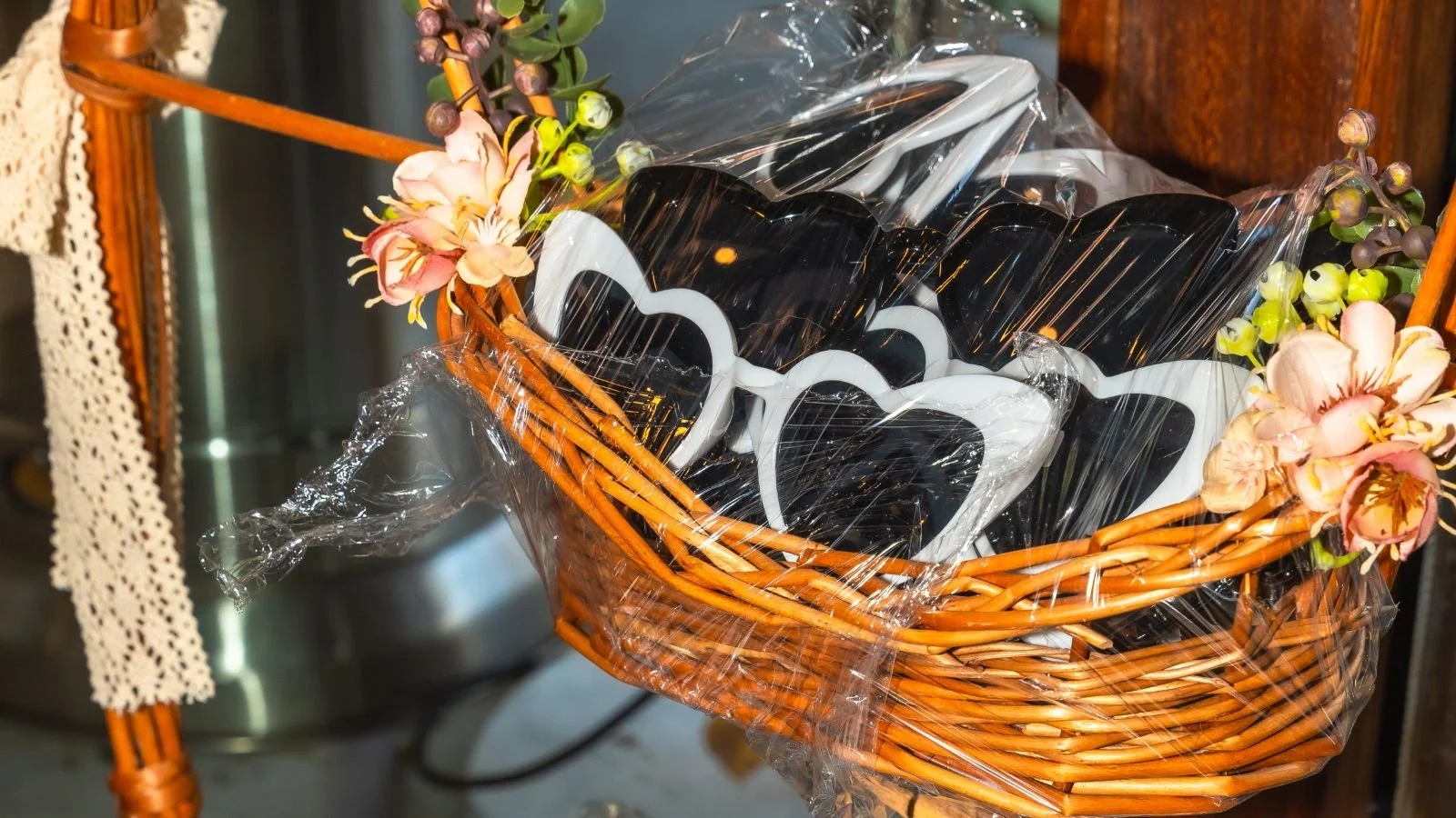 A woven basket decorated with pink and white artificial flowers, containing black and white heart-shaped sunglasses wrapped in plastic.
