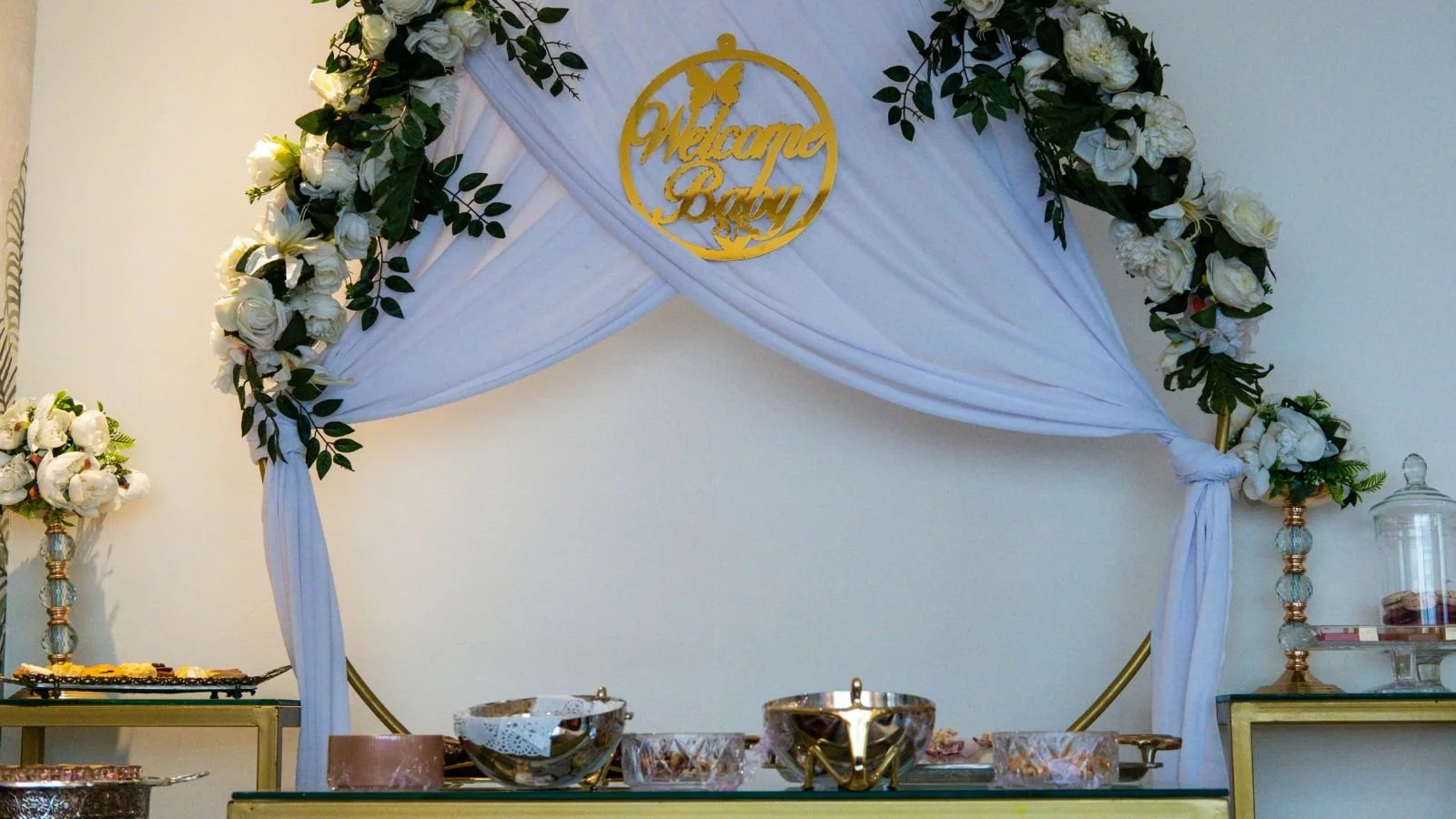 Decorative baby shower setup with a white draped fabric canopy, floral arrangements of white roses and green leaves, a gold sign that reads 'Welcome Baby,' and a table with silver bowls and sweets, along with a glass container on the side.