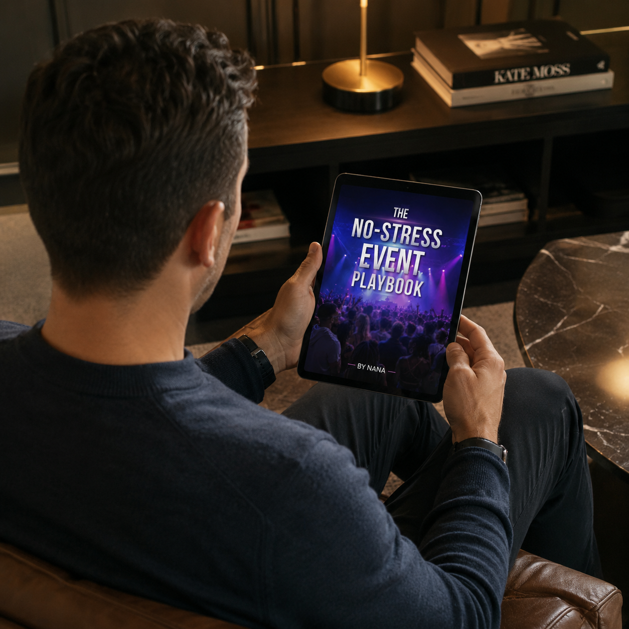 A man sitting on a brown leather couch, holding a tablet that displays the cover of a book titled 'The No-Stress Event Playbook' by Nana, with a background image of a concert crowd and stage lights.