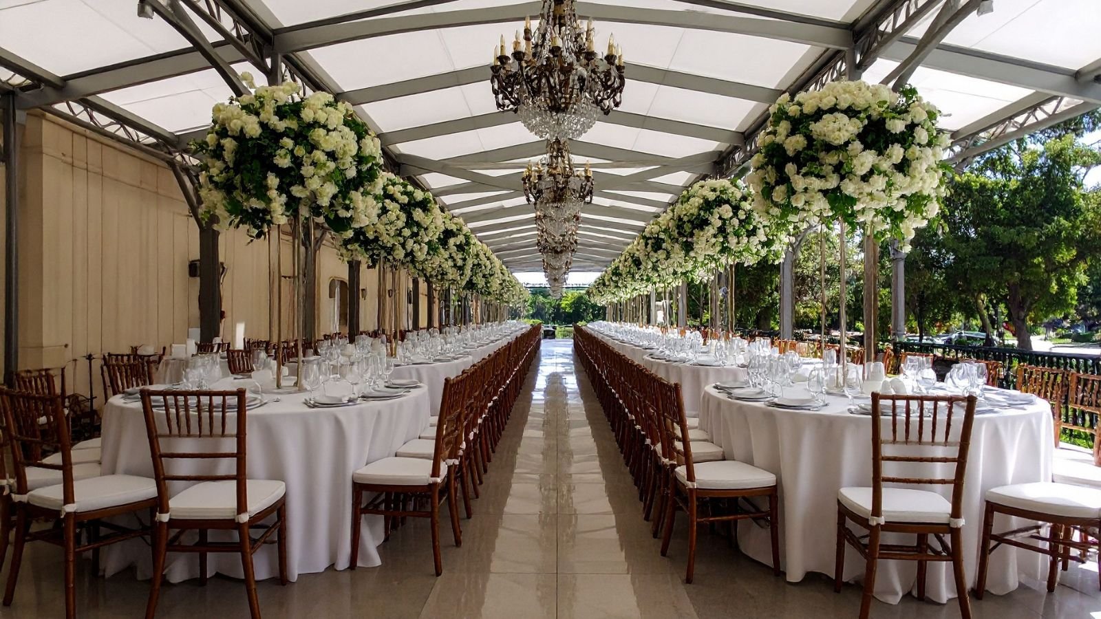 Elegant outdoor banquet setup with long tables, white tablecloths, white floral centerpieces hanging from the ceiling, chandeliers, and wooden chairs, under a transparent canopy surrounded by trees.