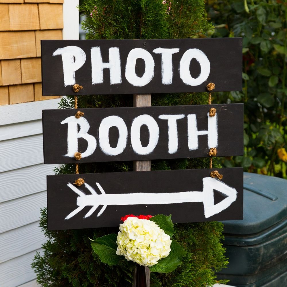 A black wooden sign with white painted letters reading 'Photo Booth' and an arrow pointing right, decorated with a white hydrangea flower and greenery in the background.