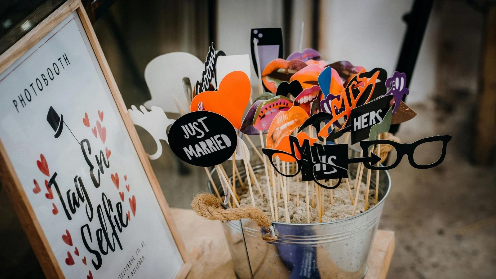 A photo booth sign and photo props on sticks in a metal bucket, including lips, glasses, hearts, and signs with phrases like "Just Married" and "Hers".
